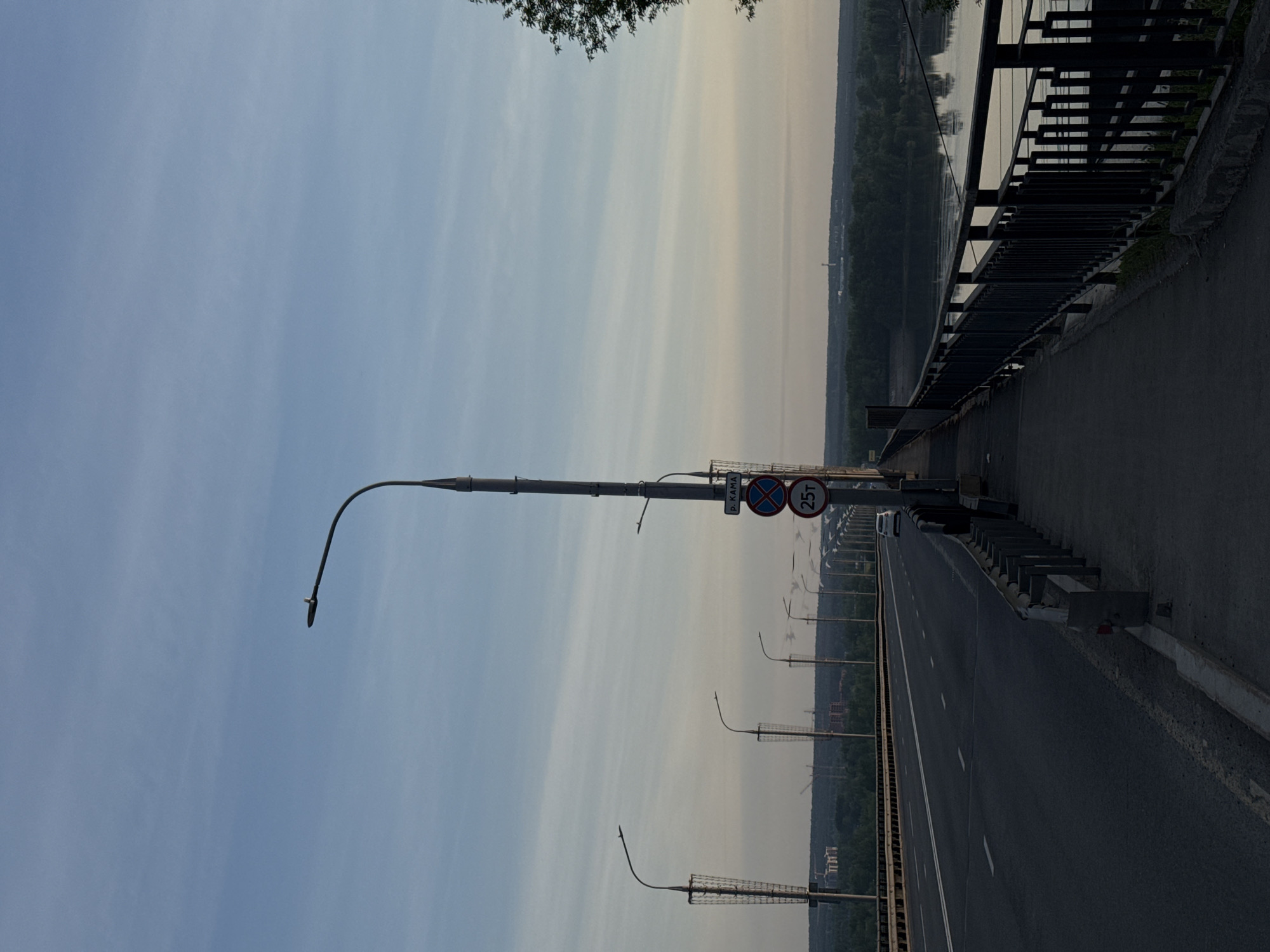 The image depicts a bridge in Perm, Russia. The photograph is taken from the bridge itself, looking down the roadway. The bridge features a metal railing on the right side and streetlights lining the road. A sign is visible with a red circle and a crossed-out symbol, along with the number "25T." A body of water is visible next to a forest. The sky is overcast. A car is seen on the bridge.