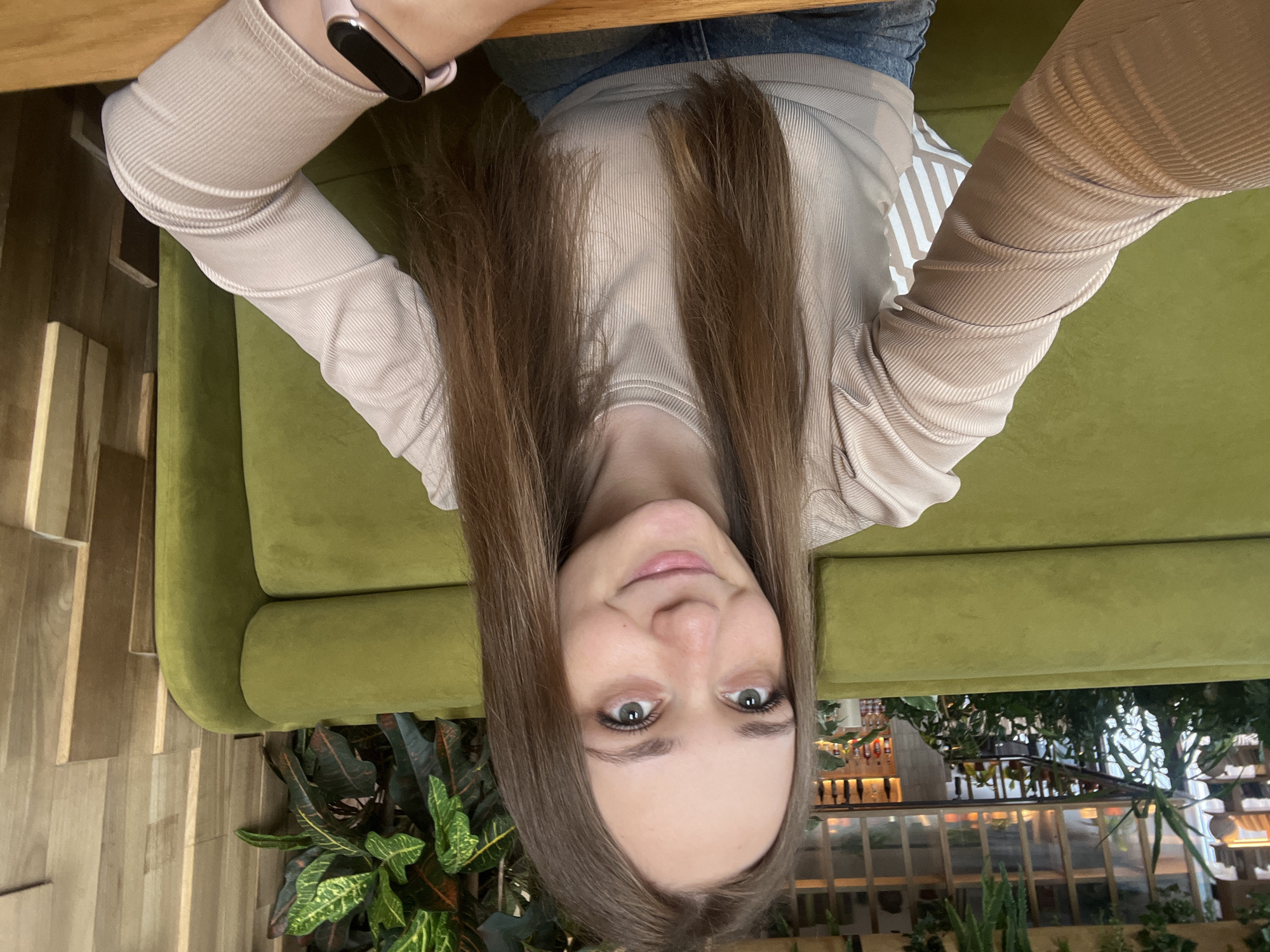 A selfie image of a young woman with long brown hair, light skin, and green eyes is rotated 180 degrees. She is wearing a light beige, ribbed long-sleeved shirt. A pink smartwatch is visible on her left wrist. She is seated on a green velvet couch in what appears to be a restaurant or lounge area. In the background, through a window, shelves of bottles and glassware can be seen, suggesting a bar setting. Plants are also visible in the foreground. Based on available information, the location may be the restaurant "Verkhnyaya Pyshma" in the city of Verkhnyaya Pyshma, Russia. The image focuses on the woman, with the surrounding environment providing context.