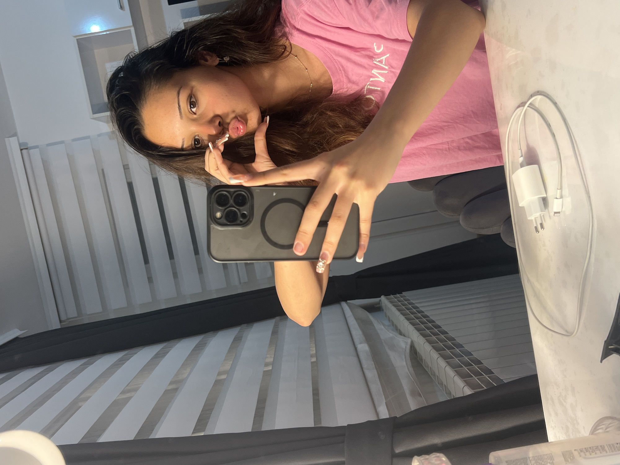 The image displays a young woman taking a mirror selfie indoors. The photograph is oriented vertically, showing the woman's head tilted towards the right. She has long, dark brown hair and is wearing a pink t-shirt with the word "SANTINAS" visible in reversed text on the front. Her expression is playful, with pursed lips and her tongue slightly sticking out. She is holding a dark-colored smartphone, likely an iPhone due to its distinctive camera array, in her left hand to capture her reflection. Her fingernails are light-colored, possibly with white tips, and her left ring finger features decorative rhinestones.
In the background, on the right side of the image, a white phone charger consisting of a wall adapter and a cable is placed on a reflective white surface, suggesting a table or counter. The left side of the background reveals white horizontal blinds or slats, possibly covering a window, and a white shelf or cubbyhole above them. A dark grey or black horizontal bar runs across the lower portion of the image, potentially part of a furniture frame or window sill. Grey upholstered seating or a headboard can be faintly seen in the mid-right background. The overall setting appears to be a well-lit interior room, possibly a bedroom or living area. The woman's pose and expression convey a casual and playful mood.