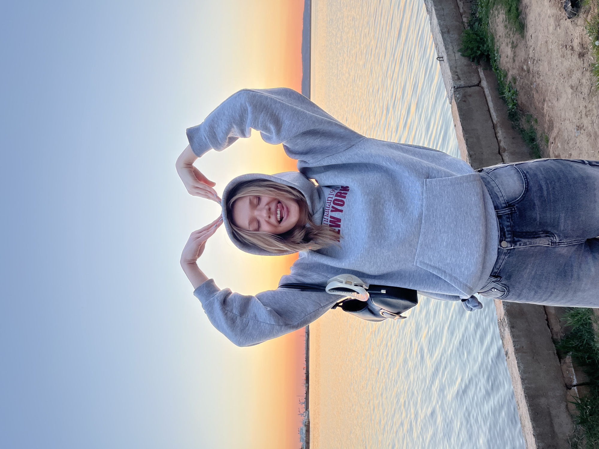 A young woman in a grey hooded sweatshirt with "NEW YORK" in red lettering and dark jeans is posing against a background of a body of water and a sunset. She is making a heart shape with her hands above her head and smiling. A black shoulder bag hangs from her shoulder. The scene appears to be outdoors, near a shoreline. Some concrete and grass are visible to the right.
The location is Chaikovsky, Russia.