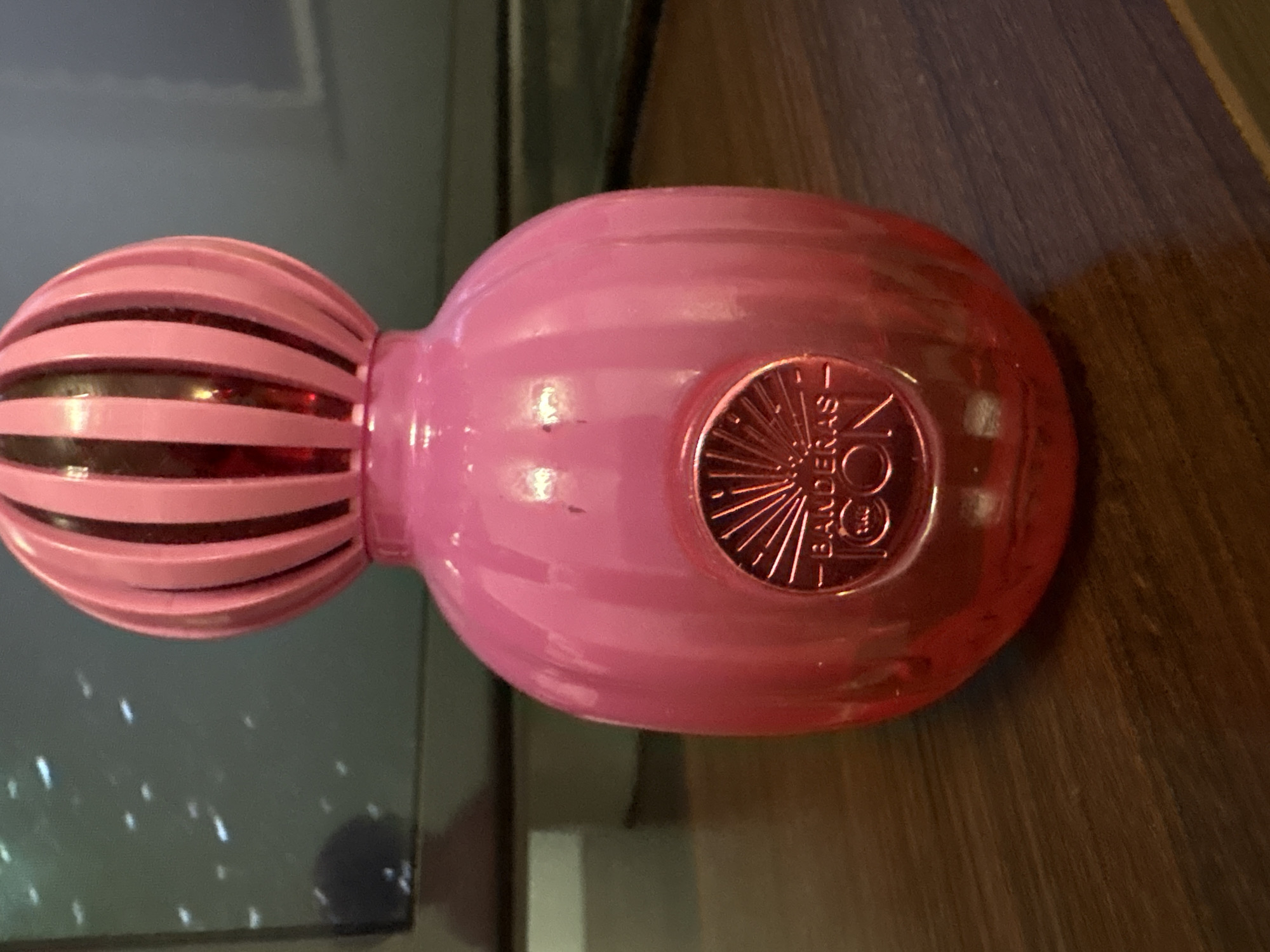 A close-up, eye-level, slightly upward-angled shot shows a pink, ribbed, globe-shaped perfume bottle with a red interior. The bottle has a round, metallic emblem on the front with the inscription "- BANDERAS ICON -". The bottle sits on a dark wooden surface. In the background, out of focus, is a dark screen, possibly a television, with some white specks and a faint reflection. There are no people visible in the image. The location in Małkowo, Poland, is not discernible from the image.