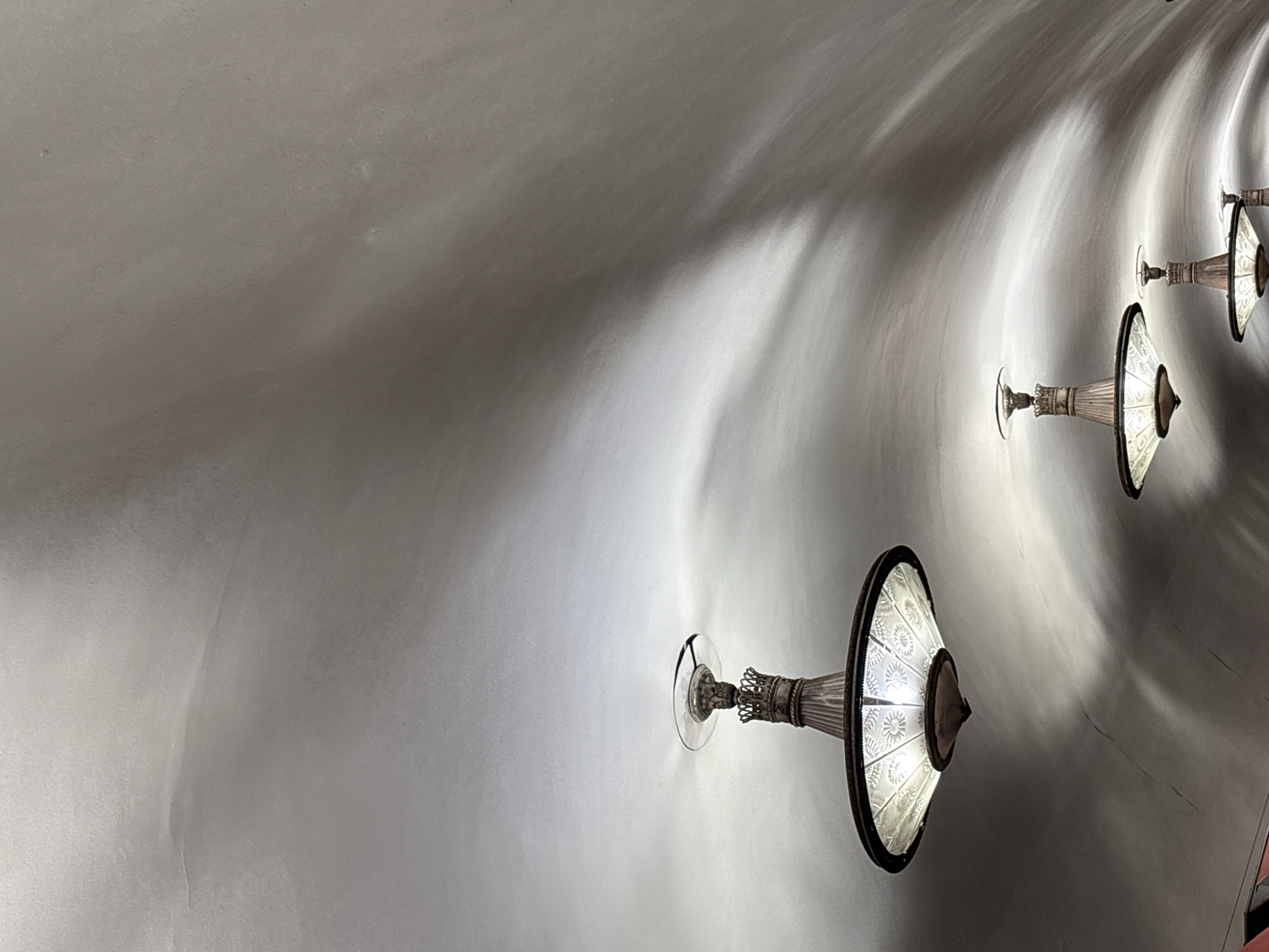 The image shows a curved, likely plastered, ceiling with three identical sconce-type lights mounted at intervals along the curve. The lights are decorative, with a glass or crystal shade patterned with designs, and a metallic base. The light from the sconces creates elongated shadows and highlights on the ceiling.
This is likely a view of a corridor within a building designed by Antoni Gaudí, specifically the Casa Batlló, located in Barcelona, Spain, not Saint Petersburg, Russia.