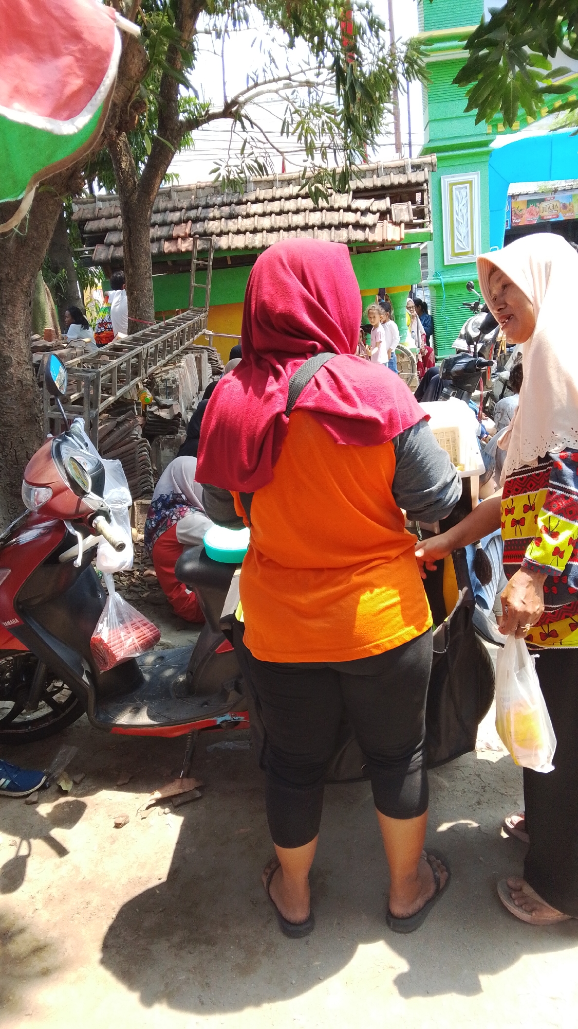 In Mancar, Indonesia, two women are engaged in conversation on a sunny day. One woman, wearing a red hijab and an orange shirt, is facing away from the camera, holding her stomach. The other woman, in a light-colored hijab and a colorful shirt, faces the first woman and holds a plastic bag of groceries. They are standing next to a red motorcycle parked on a dirt ground. In the background, there are buildings, trees, and other people, suggesting a bustling outdoor market or street scene.
