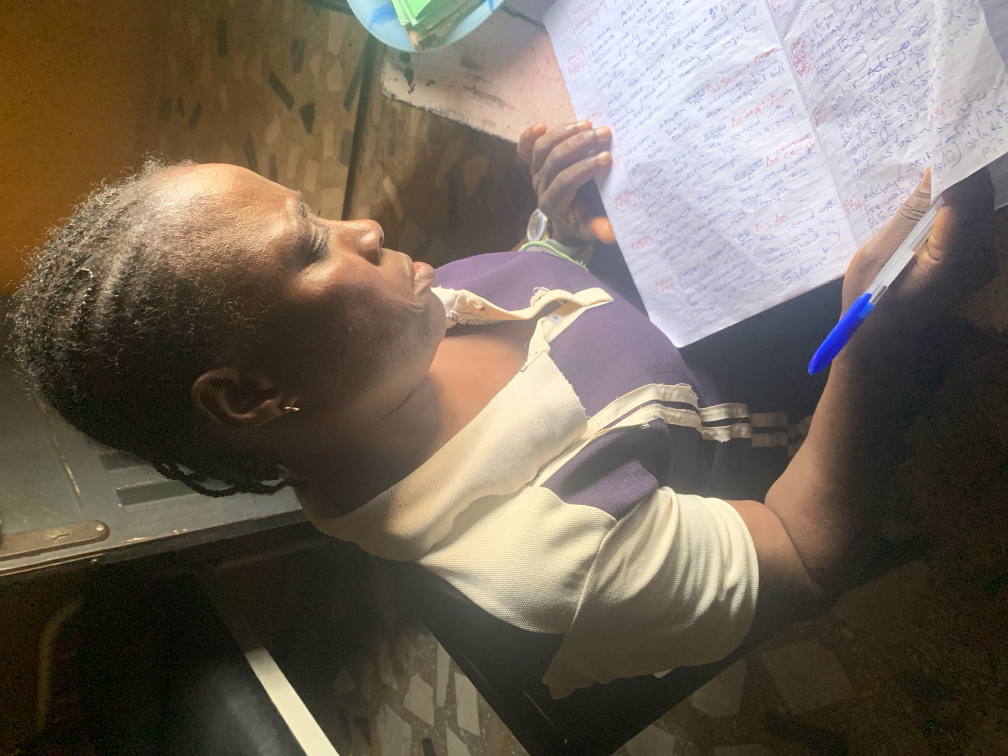An overhead, medium shot captures a Black woman seated at a table, engrossed in reading and writing in a notebook. She is wearing a purple and cream collared shirt and has her hair in braided cornrows. A blue pen is in her right hand, poised over the notebook. The background shows a dimly lit room with a wooden door frame visible on the left and a yellow-orange wall with a tiled pattern on the right. The scene appears to be an interior shot within a residential setting in Shagamu, Nigeria.