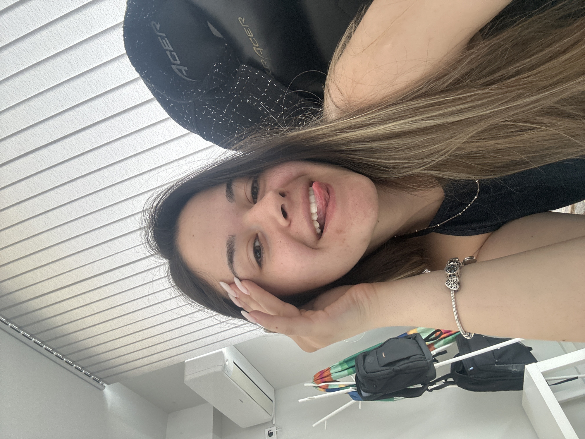 A woman is in a room in Sofia, Bulgaria. She is sitting in an Acer gaming chair. She is smiling and sticking out her tongue. She has long brown hair and is wearing a black top and a bracelet with charms. The room has white walls and a white ceiling with horizontal blinds. An air conditioning unit is mounted on the wall. A drying rack with black bags is in the background.