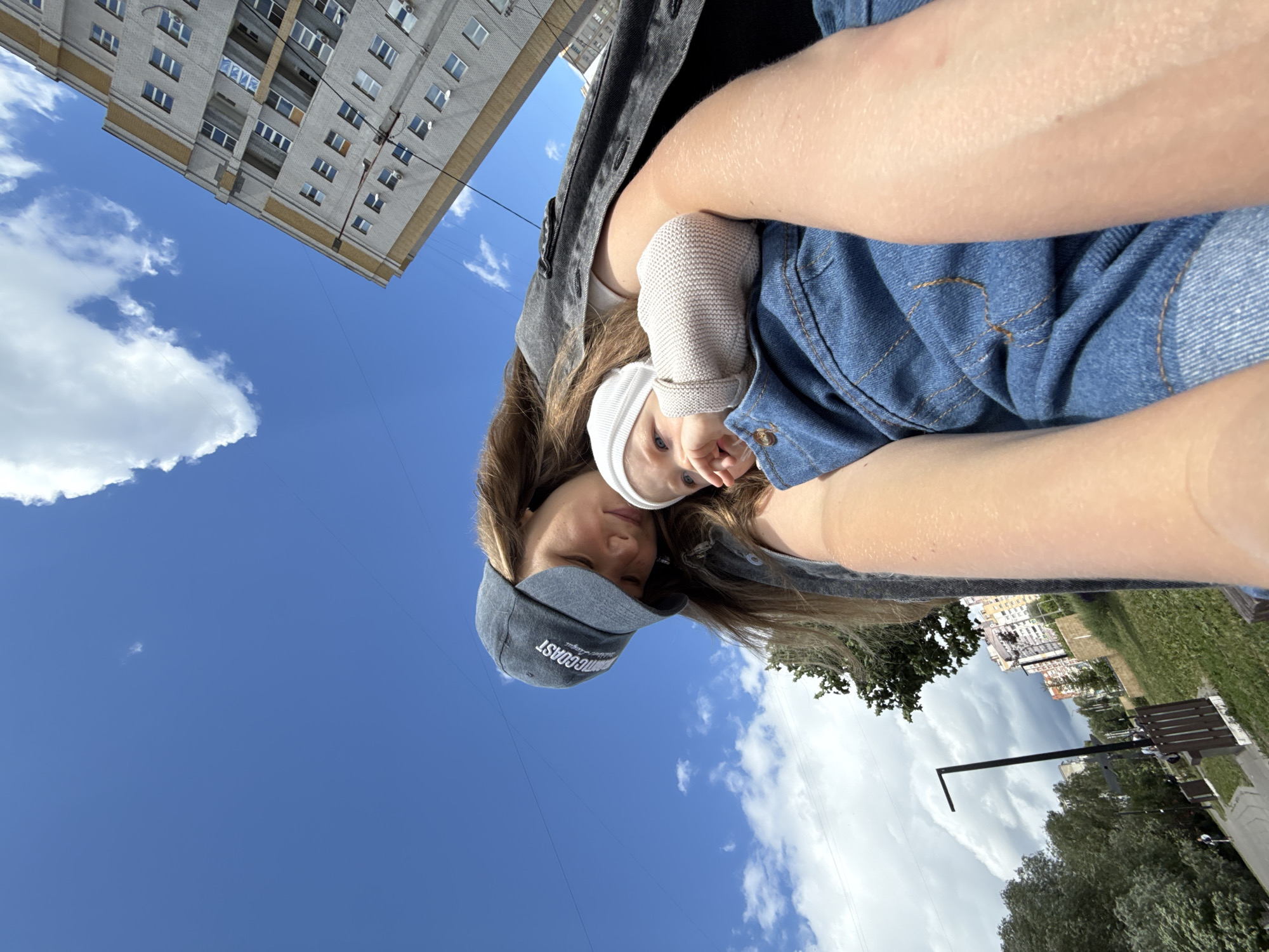 This is a selfie taken outdoors, likely during the daytime, as indicated by the bright blue sky and visible sunlight. The photographer appears to be a young woman, holding a baby wrapped in a white beanie and a grey knitted garment. Both are wearing casual clothing, with the photographer in a denim jacket and shorts, and the baby in a beanie. The setting is a park or a public outdoor area, with a grassy area, trees, a paved walkway, and park benches visible in the background. Further in the distance, there is a multi-story apartment building, typical of urban environments in Vladimir, Russia. The sky is a vibrant blue with scattered white clouds, suggesting fair weather. The overall mood appears relaxed and casual, with the photographer looking towards the camera and the baby appearing to be looking away. There is text visible on the photographer's baseball cap, which reads "STORMCOAST". The image is oriented with the photographer and baby at the top, and the ground and buildings below, suggesting the photo was taken looking upwards.
