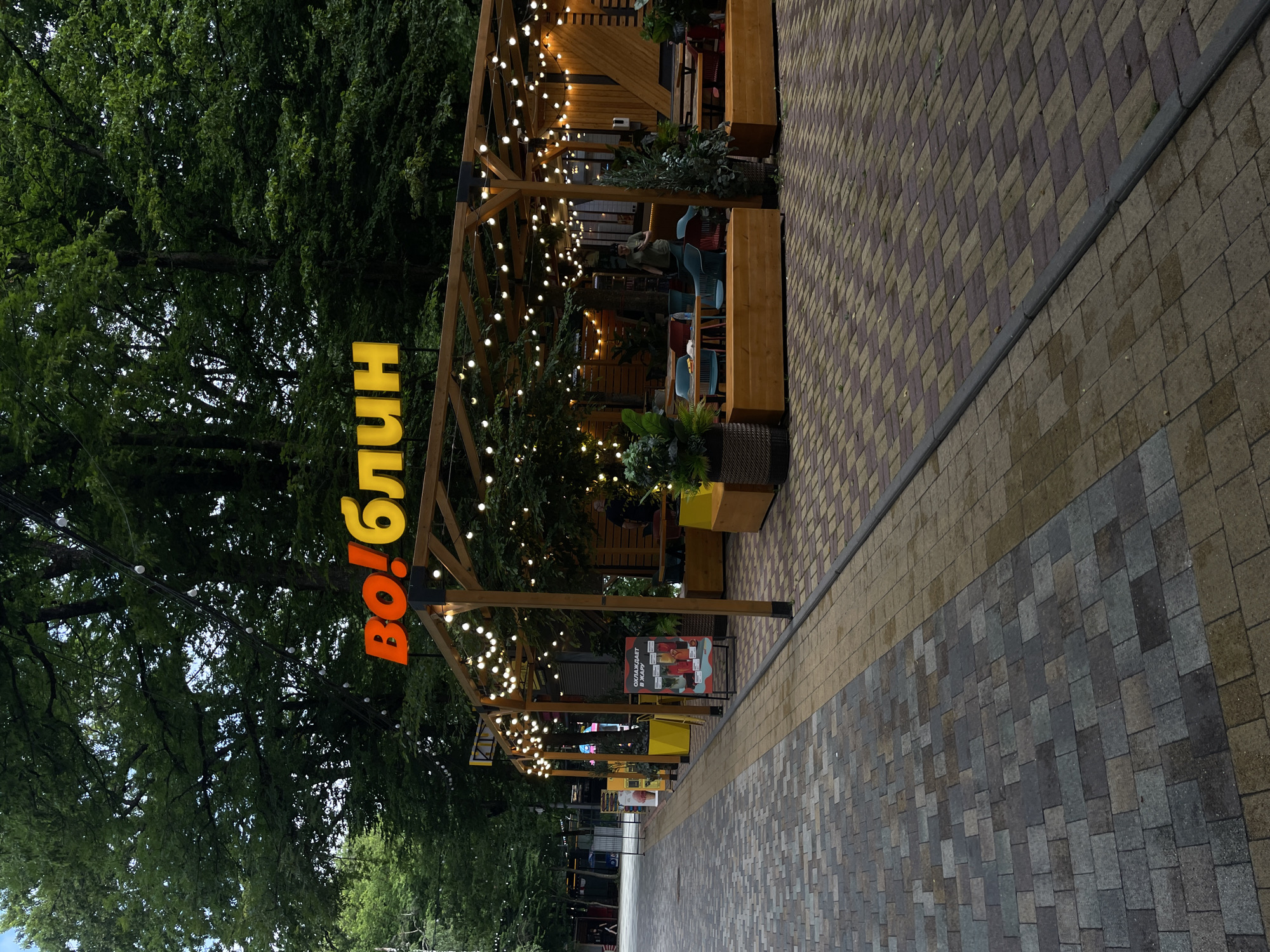 The image depicts an outdoor dining area under a wooden pergola adorned with string lights. A brightly colored sign with Russian text, "во!БЛИН," is prominently displayed at the top of the structure. The area features wooden tables and chairs, as well as planters with greenery. In the background, there are trees and other buildings, suggesting an urban environment. The ground is paved with interlocking bricks of various colors. A stand with a menu is visible to the left, and a few people are present in the seating area. This scene is likely a cafe or restaurant in Stavropol, Russia, given the presence of the Russian language on the sign and the overall aesthetic.