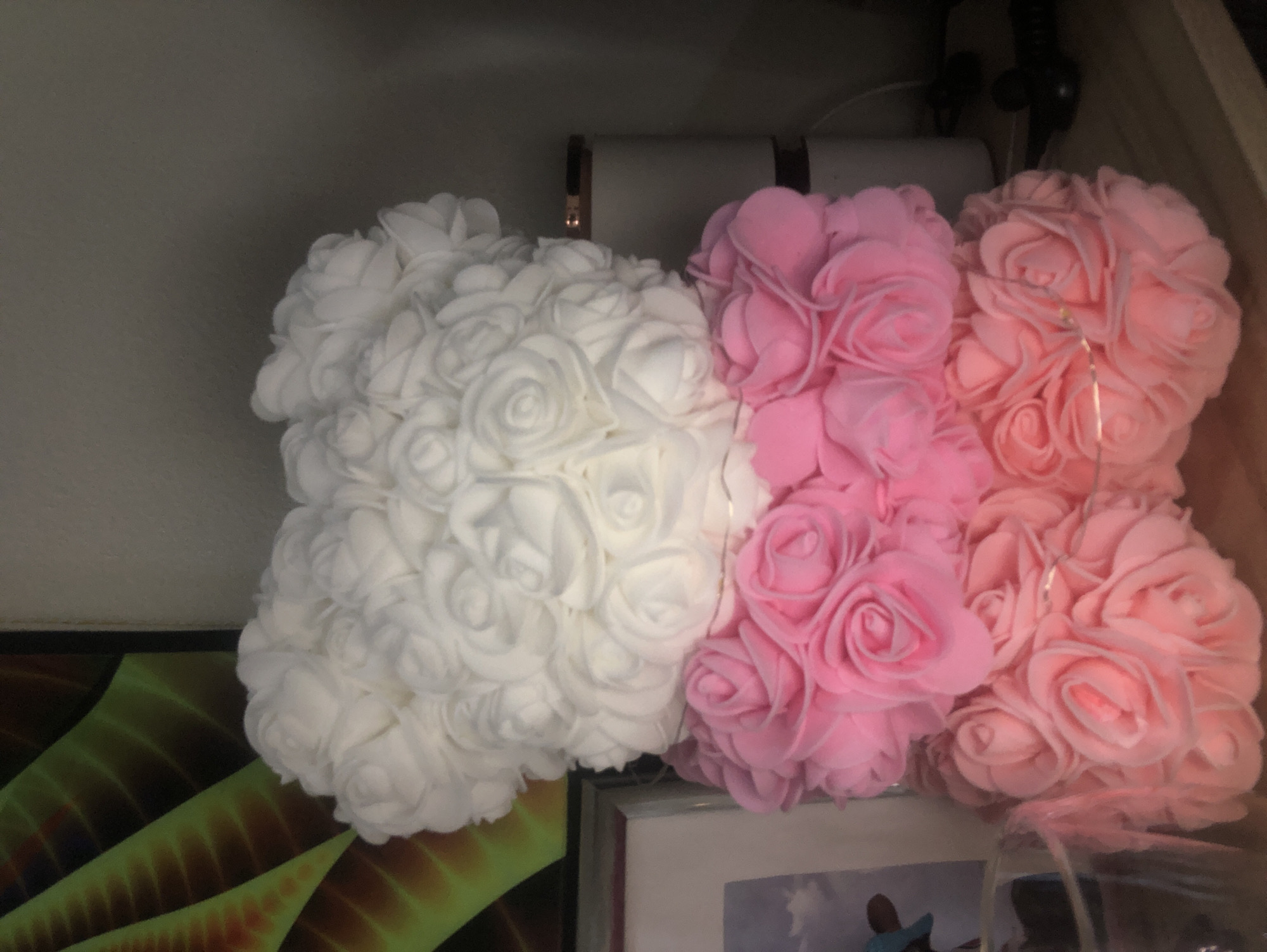Two plush-like forms, crafted from numerous artificial roses, are displayed indoors in what appears to be Baan Chaloklum, Thailand. On the left, one large object is entirely composed of white roses, while to its right, two similar objects are joined, made of light pink and slightly deeper pink roses. Delicate string lights are subtly woven into the pink rose arrangements, suggesting they are decorative items, possibly gifts or home décor.
The objects are positioned on a surface, likely a shelf or counter. In the background, above the roses, indistinct items such as tubes or rolls and some wires are visible on another shelf, suggesting a storage or display area. Below the rose arrangements, to the left, a vibrant, abstract pattern in shades of green and orange is partially visible. Directly beneath the pink roses, a white picture frame containing an image (details unclear) is partially shown. No human subjects are present, and no specific activity is taking place beyond the static display of these decorative items. The indoor setting implies artificial lighting is the primary light source, and there is no indication of weather. No visible text is present in the image.