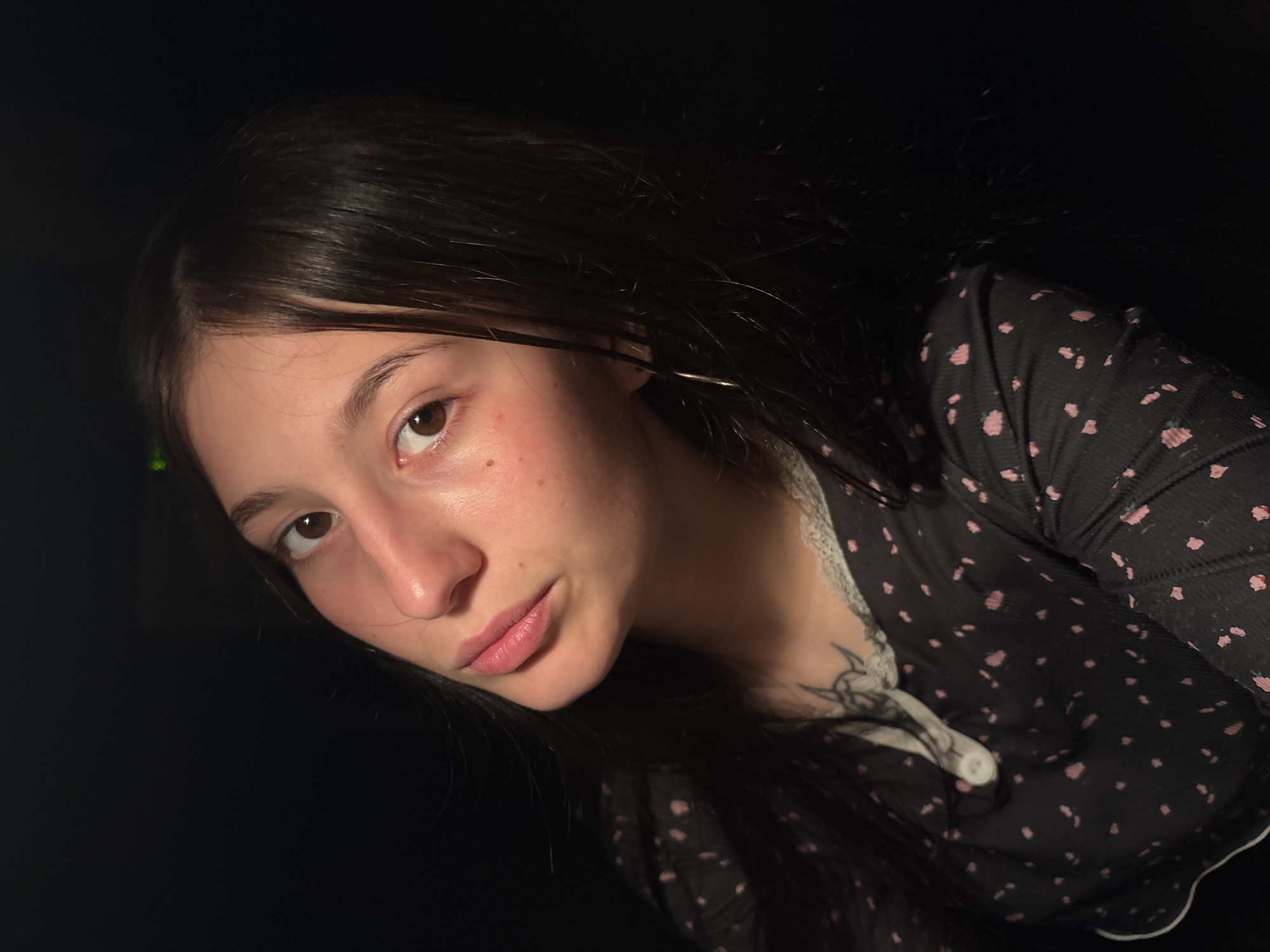 A close-up portrait features a young woman with long, dark hair and brown eyes, looking directly at the viewer with a neutral, possibly pensive expression. She has a mole below her right eye and appears to have fair skin with some light blemishes or freckles. Her lips are a natural light pink color.
She is wearing a dark, possibly black, long-sleeved top adorned with small pink polka dots or floral patterns. The top features a white lace trim along the neckline and a partial white button placket with one visible white button. A dark, possibly tribal or abstract, tattoo in black ink is visible on her upper chest/shoulder area, partially covered by her clothing. A silver-colored hoop earring is visible in her right ear.
The background is almost entirely dark, suggesting the photograph was taken in a dimly lit environment, possibly indoors at night or evening. Lighting appears to be directed from the front or side, illuminating her face and upper body while leaving the background in shadow. There's a subtle greenish glow or reflection near the top left of her head. The image is framed at an angle, with her head tilted slightly, giving the impression of a close, intimate shot. No text is visible in the image. The location context provided is Фоки, Russia.