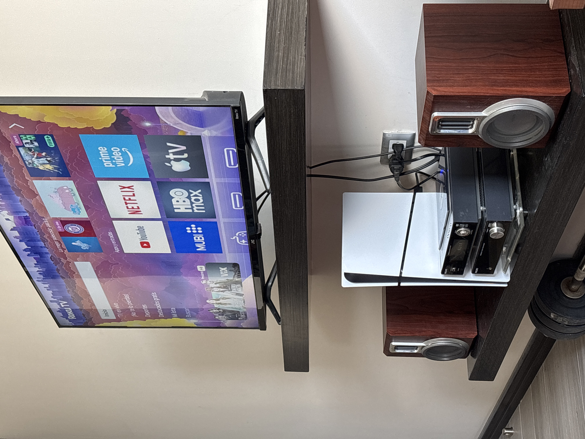 Here is a description of the image.
The image shows an entertainment center set against a light beige wall. A flat-screen television, displaying the Roku TV interface with icons for various streaming services like Netflix, YouTube, Prime Video, and Apple TV, is mounted on the wall. The television's bezel is black.
To the right of the television, a dark wood-grain shelf unit is visible. On one of the shelves rests a white PlayStation 5 console and a smaller black electronic device. Above and below the console are dark wood-grain speakers with silver-colored circular fronts. Wires and cables connect these devices.
In the lower right of the image, a glimpse of a weight or dumbbell is visible. The scene is well-lit, suggesting an indoor environment. The overall impression is of a modern entertainment setup within a home.