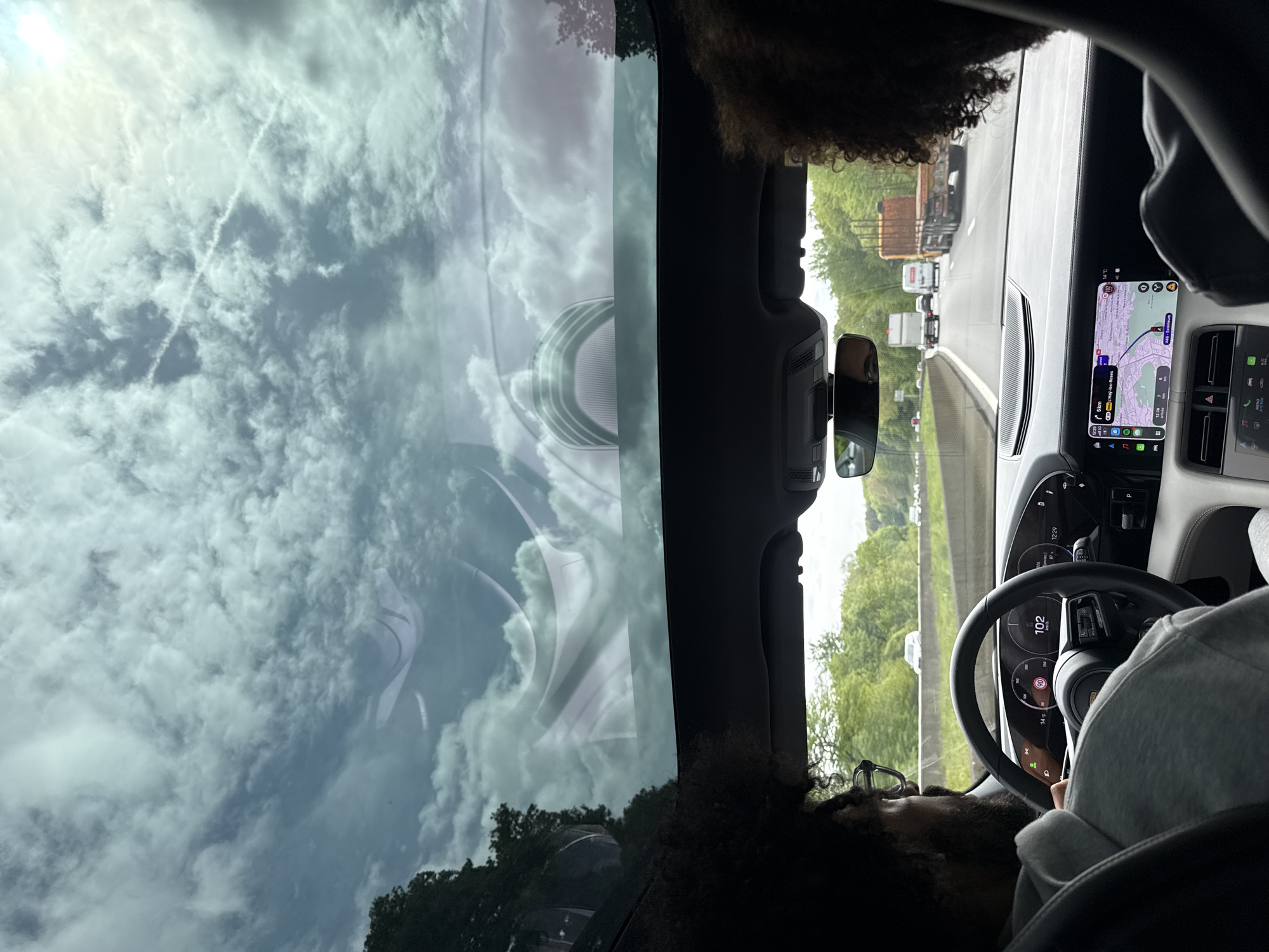 The image shows a view from inside a car, looking out through the windshield and the driver's side window. The car appears to be on a highway with traffic congestion, as several vehicles are visible ahead. The driver, partially visible, is holding the steering wheel, and the dashboard instruments indicate the car is in motion. The navigation system is displaying a route map, with the destination labeled as Châtenay-Malabry, France. The sky is cloudy, and the reflections are visible on the windshield. The overall environment suggests daytime travel.