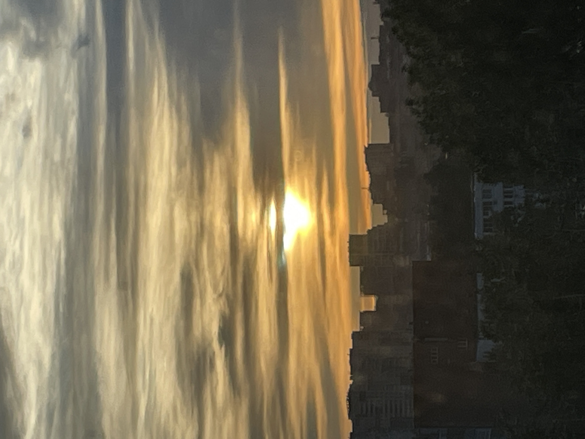 The image depicts a cityscape at sunset or sunrise in Izhevsk, Russia. The sun is visible through a break in the clouds, which are layered and streaked with gold and gray hues. To the right of the clouds, buildings are silhouetted against the bright sky. Trees and greenery are visible in the foreground. The buildings appear to be residential and commercial, consistent with an urban environment.