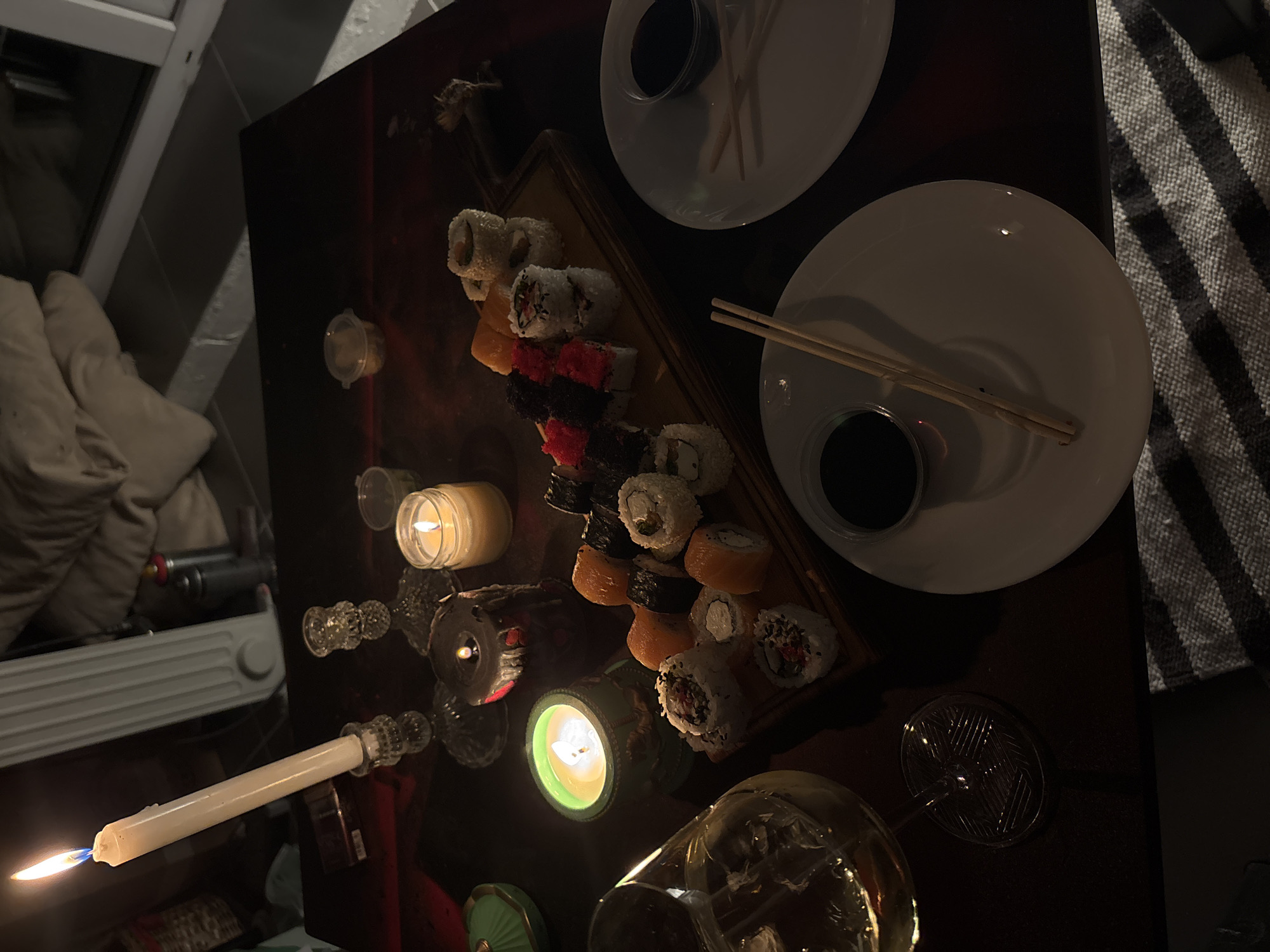 The media file captures an intimate and dimly lit indoor scene in Krasnoyarsk, Russia, featuring a sushi dinner set for two. The primary subjects are various types of sushi rolls artfully arranged on a wooden serving board, including uramaki with white sesame seeds, red roe, and black roe.
Two white plates are visible on the dark, reflective table surface, each accompanied by a pair of wooden chopsticks and a small bowl filled with dark soy sauce. A clear glass containing a drink is also present. The scene is illuminated almost entirely by several lit candles, creating a warm and cozy ambiance indicative of nighttime or a late evening event. One tall, white taper candle, a cream-colored jar candle, and other smaller decorative candles with green and dark holders cast soft, flickering light, reflecting on the polished table.
In the background, partial views of household items include a textured, light-colored fabric (possibly a blanket or cushion) on the left, a white object resembling a heater or part of a window frame, and a striped black and white textile on the right side of the table. No people are visible. The setting evokes a sense of relaxation, perhaps a romantic dinner or a quiet evening at home. No discernible text is visible in the image.