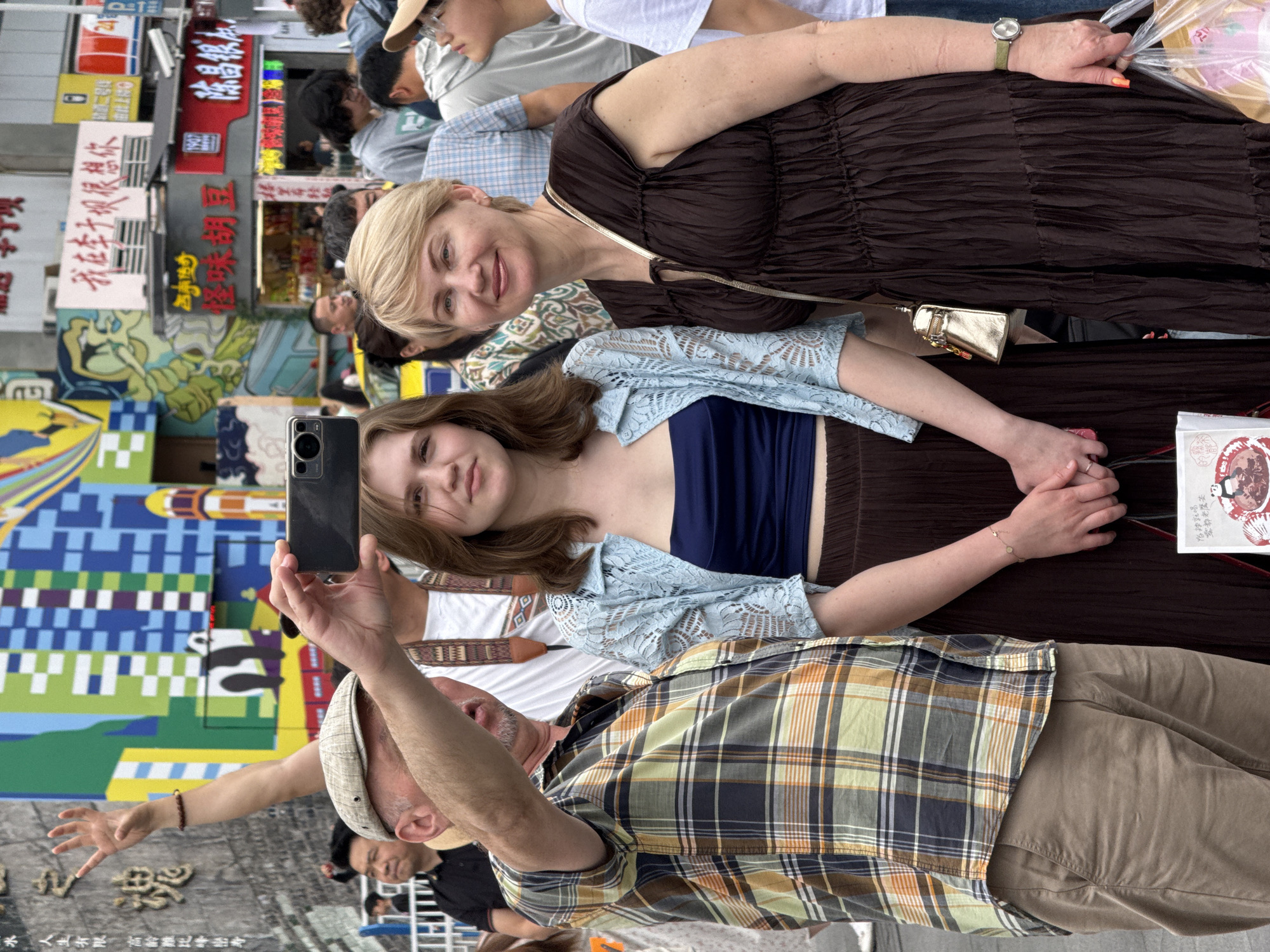 A group of three people, two women and one man, are captured in a lively outdoor setting, likely a street or market in Yuzhong District, China. The image is rotated 90 degrees clockwise.
In the foreground (bottom right of the rotated image), a man with a light-colored plaid shirt, tan pants, and a flat cap is holding a black smartphone, seemingly taking a picture or video. His left arm is extended, with his hand making a peace sign.
To his left (center of the rotated image), a younger woman with brown hair wears a dark blue tube top and a light blue, lacy long-sleeved cardigan. She is looking directly towards the smartphone with a soft expression. Her hands are clasped.
Behind her (top left of the rotated image), an older blonde woman smiles towards the camera. She is dressed in a dark brown, ruffled maxi dress and wears a watch with a green strap on her left wrist. A small golden purse hangs from her shoulder, and she holds a clear plastic bag in her right hand.
The background is bustling with activity and colorful details. Various market stalls and shops line the street, adorned with numerous Chinese signs. A large, brightly colored mural with geometric patterns and stylized figures is visible on a wall. Other people are partially visible in the background, contributing to the busy atmosphere.
Notable text from the signs includes: "水人生有限,高於難比峰巒寿" (Life is limited, surpassing difficult peaks like mountains), "我在本于现很想你" (I really miss you now), "BRING", "陈昌银" (Chen Changyin, likely a brand or name), and "俗辣就喝 雲都老鷹茶" (Colloquially spicy, just drink Yundu Old Eagle Tea). The scene appears to be taking place during the daytime, under bright, clear weather conditions. The overall mood conveyed by the subjects' expressions is pleasant and relaxed.