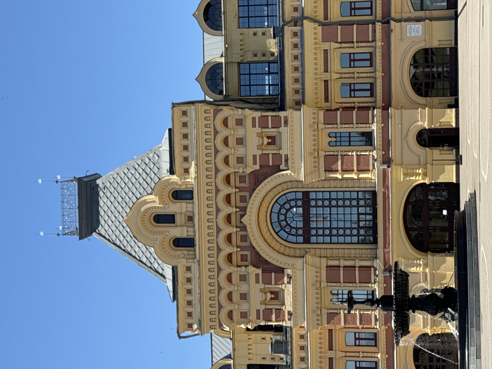 The image is a vertical shot of a grand, ornate building with a clear blue sky in the background. The architecture is detailed, featuring yellow and reddish-brown brickwork, decorative moldings, and numerous windows, some of which are arched and have intricate geometric patterns. The building has a steeply pitched, grey tiled roof with a pointed turret-like structure at one end, topped with metalwork.
A black, multi-tiered fountain with water cascading down is prominently positioned in the foreground, partially obscuring the lower part of the building. The fountain appears to be made of dark metal and has a classic, sculptural design. The sunlight suggests it is daytime, and the clear sky indicates good weather.
The perspective is from a low angle, looking up at the building and the sky. The composition emphasizes the architectural grandeur of the building and the elegance of the fountain. There are no people visible in the frame. The only visible text is a series of repeating square shapes that resemble stylized letters or symbols, forming a decorative frieze on the building's facade. The location is identified as Nizhny Novgorod, Russia.