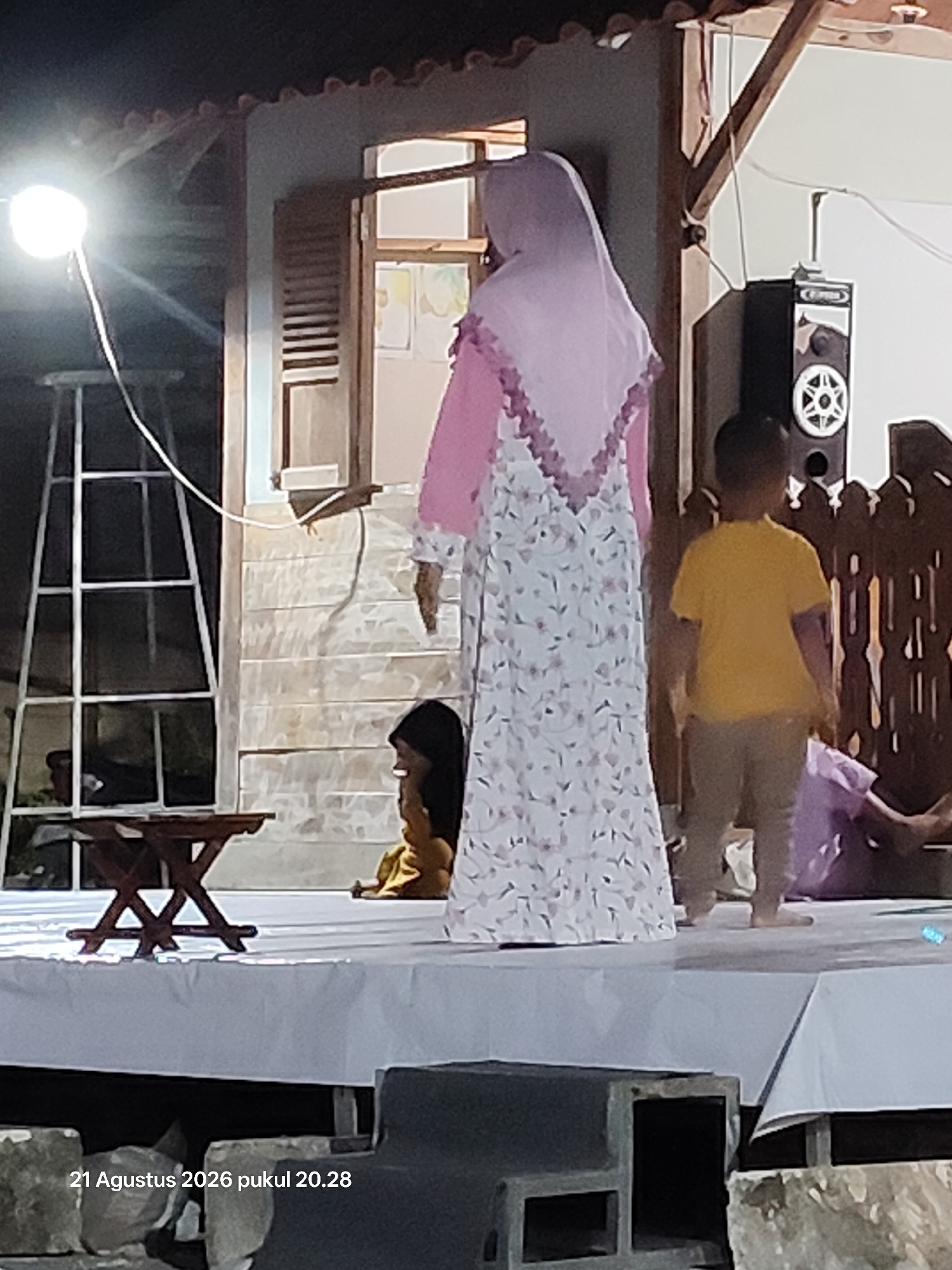 The image depicts an outdoor evening event in the city of Wedi, Indonesia. A stage is set up, covered with a white cloth. On the stage, a woman wearing a pink hijab and a floral dress stands prominently. To her right, a young boy in a yellow t-shirt and light-colored pants is visible from behind, and further to the right, another person is partially visible wearing a purple garment. To the left of the woman, a small wooden table is placed on the stage. In the background, there is a building with open shutters and a speaker mounted on it. A bright light is illuminating the scene from the left, creating a spotlight effect. A date and time stamp, "21 Agustus 2026 pukul 20.28", is visible in the bottom left corner of the image.