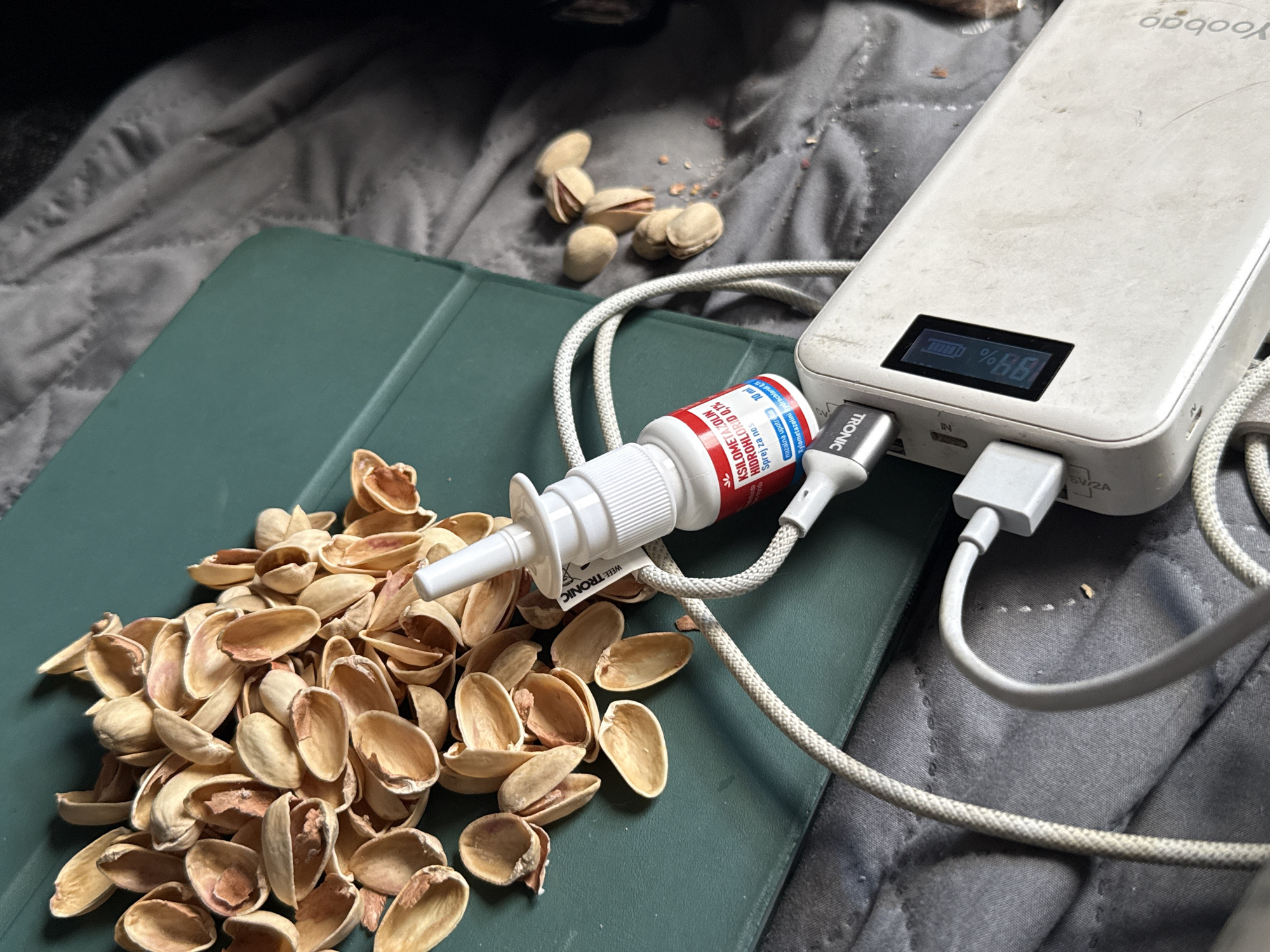 This close-up, medium-shot image depicts a collection of pistachio shells scattered on a dark green, tablet-like surface. The shells are light brown and vary in size and shape, with some appearing more broken than others. A small, white nasal spray bottle with a red and blue label is positioned amongst the shells, its nozzle pointing towards the left. A white cord is attached to the spray bottle and runs towards a white power bank, which is also partially visible in the frame. The power bank has a digital display showing "88%" and a battery icon. Another white cord is plugged into the power bank. The background is a dark gray, quilted fabric, possibly a blanket or bedspread. A few whole pistachios are visible further up and to the left of the frame. The lighting appears to be artificial and casts shadows. The scene is set indoors, as indicated by the power bank and the fabric background. There are no people visible in the image. The overall impression is one of a still life arrangement, possibly suggesting a moment of rest or break, with the nasal spray and charging device hinting at personal use. The brand "TRONIC" is visible on the power bank and the spray bottle's label. Text on the spray bottle is partially legible, including "KSILOMETAZOLIN HIDROHLORID 0,1% Sprej za nos". The location context is Kanjoši, Montenegro.