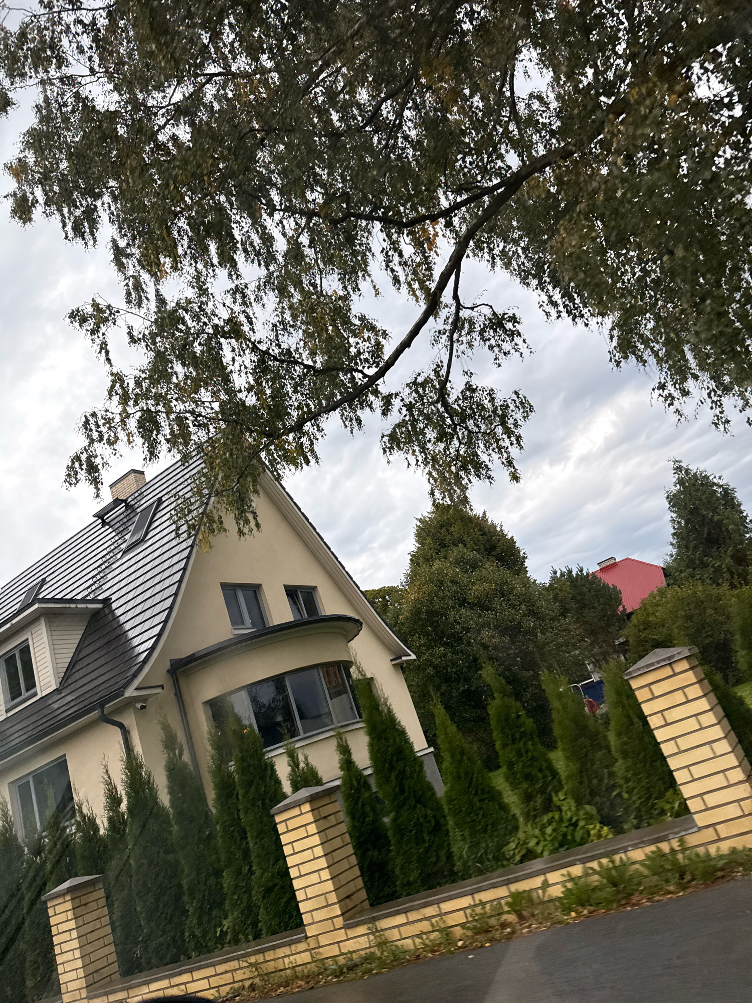 The image depicts a street view in Tallinn, Estonia. In the foreground, a low beige brick wall with pillars lines the street. Behind the wall, a row of tall, dark green evergreen trees stands in front of a light yellow house. The house has a dark gray, multi-pitched roof with dormer windows and a prominent chimney. To the right of the house, a bright red-roofed building is visible amongst more trees. Overhanging the scene from the top is a large tree with dark green leaves. The sky is overcast with gray clouds.