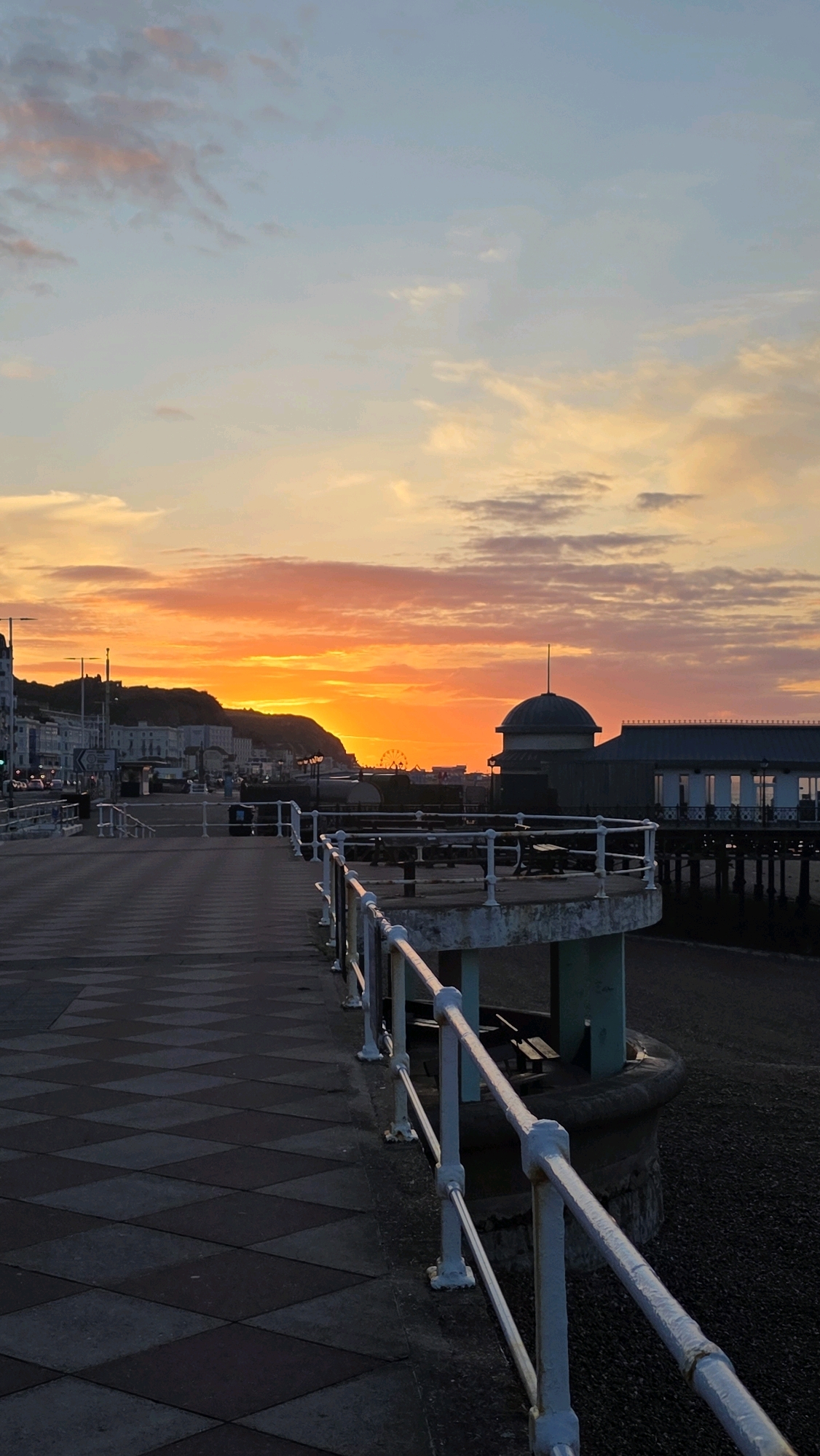 The image depicts a sunset over a pier in St Leonards, United Kingdom. A paved walkway with a diamond pattern extends into the foreground, bordered by a white metal railing. Beyond the walkway, a section of the pier features a circular structure with a domed roof and a building with a long, flat roof. In the distance, a Ferris wheel is visible against the colorful sunset sky, with a hillside and town buildings silhouetted behind it. The sky displays a gradient of colors, from pale blue at the top to vibrant oranges and yellows near the horizon. The beach area below the pier is covered in dark pebbles. There are no people clearly visible in the frame.