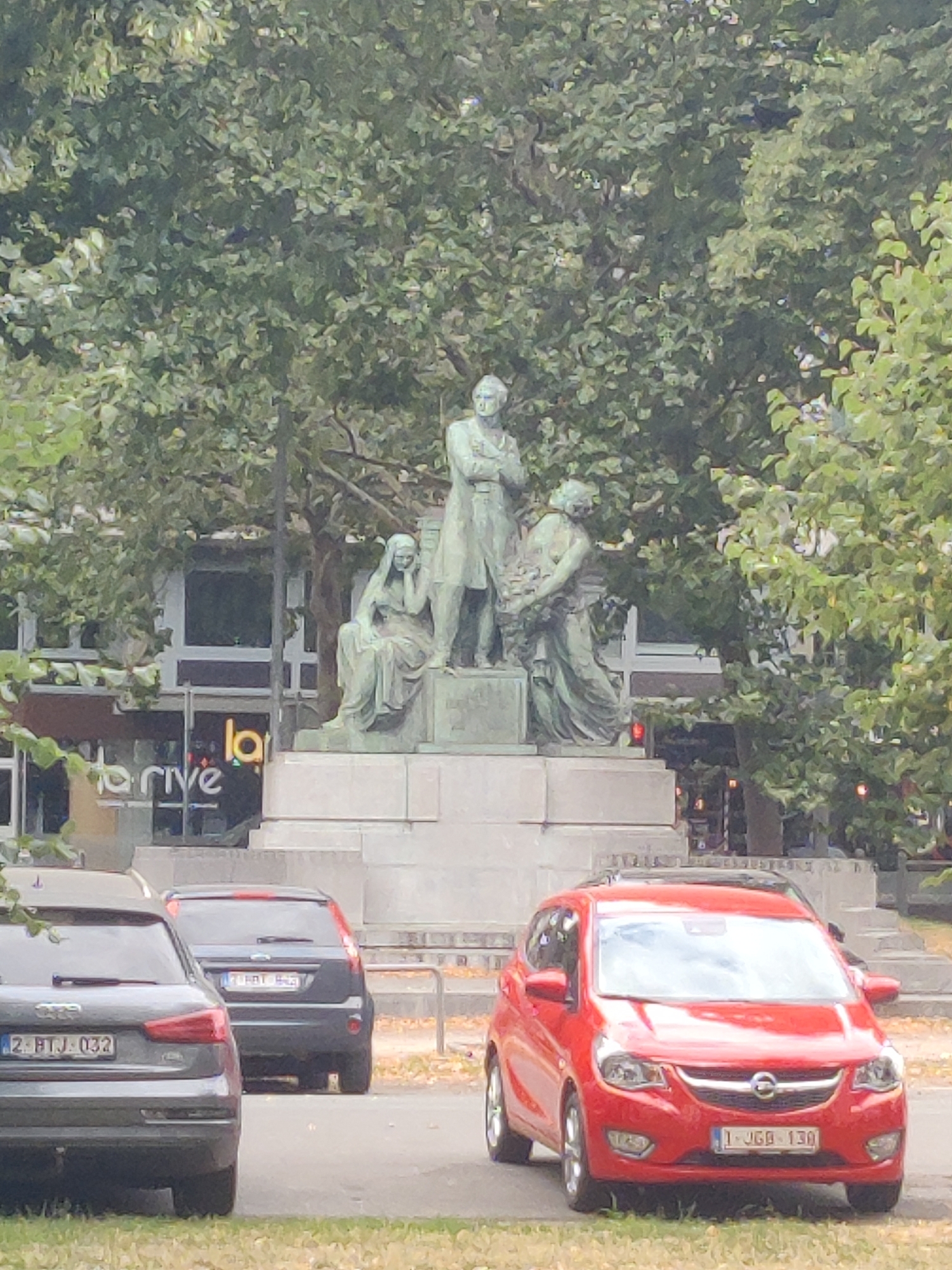 The image shows a statue in a park-like setting with trees in the background. In the foreground, there are several parked cars, including a red Opel Karl. Behind the cars, a statue is visible, depicting three figures. The central figure is a standing man in a suit, with two other figures seated or kneeling beside him. The statue is on a stone pedestal. In the background, a building with large windows and a sign that reads "drive" is partially visible. The scene appears to be in an urban environment, likely a public square or park in Liège, Belgium, given the license plates on the cars.