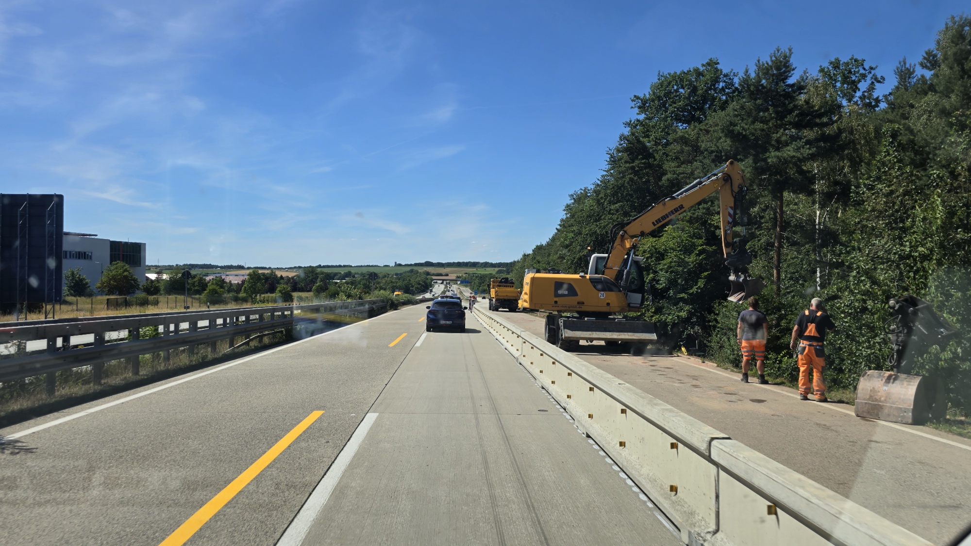 The image captures a construction site on a highway, likely in or near Kaiserslautern, Germany. The scene is dominated by a yellow Liebherr excavator positioned on the right side of the road. Two construction workers, both wearing orange high-visibility trousers and dark tops, are present near the excavator. One worker is facing away from the camera, while the other is partially obscured by the excavator. A large metal bucket lies on the ground next to them.
The highway has multiple lanes, with traffic moving in the distance. A dark-colored car is traveling in one of the lanes, and a yellow dump truck is also visible. The road is marked with yellow and white lines. On the left side of the highway, a metal guardrail separates the road from an embankment with vegetation and a large industrial-looking building in the background. The sky is clear and blue, with some wispy clouds. The surrounding environment is characterized by a mix of trees and fields, suggesting a semi-rural or suburban setting. The overall impression is one of ongoing road construction or maintenance work.