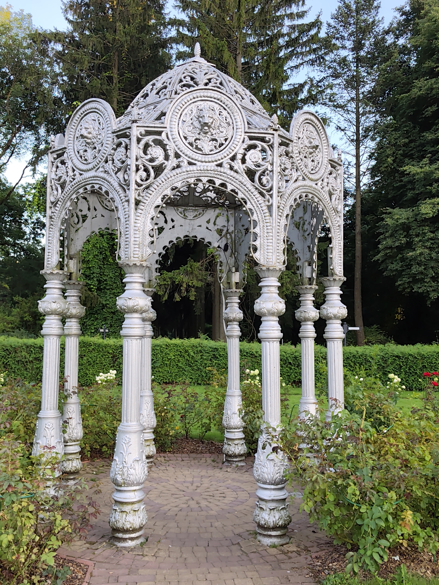 This is a photograph of a decorative white cast-iron gazebo located in Minsk, Belarus. The gazebo features intricate metalwork, including ornate columns, arches, and a domed roof with decorative circular motifs. The gazebo is situated outdoors in a garden setting, surrounded by rose bushes and a neatly trimmed hedge. In the background, tall evergreen trees are visible against a clear blue sky. The ground within and around the gazebo is paved with brick in a circular pattern. There are no people present in the image.