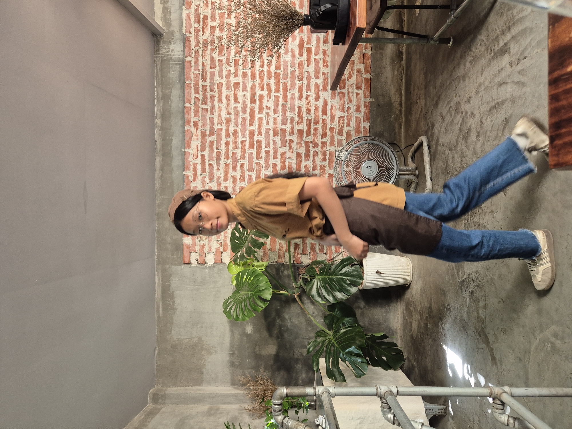 The image depicts an indoor scene, likely a cafe or restaurant, with a rustic, industrial aesthetic. The walls are a mix of exposed brick and concrete. On the left, a large Monstera plant in a white pot stands near a metal pipe fixture. The center of the image features a young woman wearing glasses, a tan cap, a tan shirt, a brown apron, and blue jeans. She is walking to the right, looking towards the camera. Behind her, a white electric fan sits on the floor in front of the brick wall. To the right, a wooden table and chairs are visible, and a vase of dried flowers sits on a black tray on the table. The floor is concrete. The lighting suggests it is daytime, with sunlight casting shadows on the floor. While the image is set in Da Nang, Vietnam, this detail is not visually inferable from the provided photograph itself.