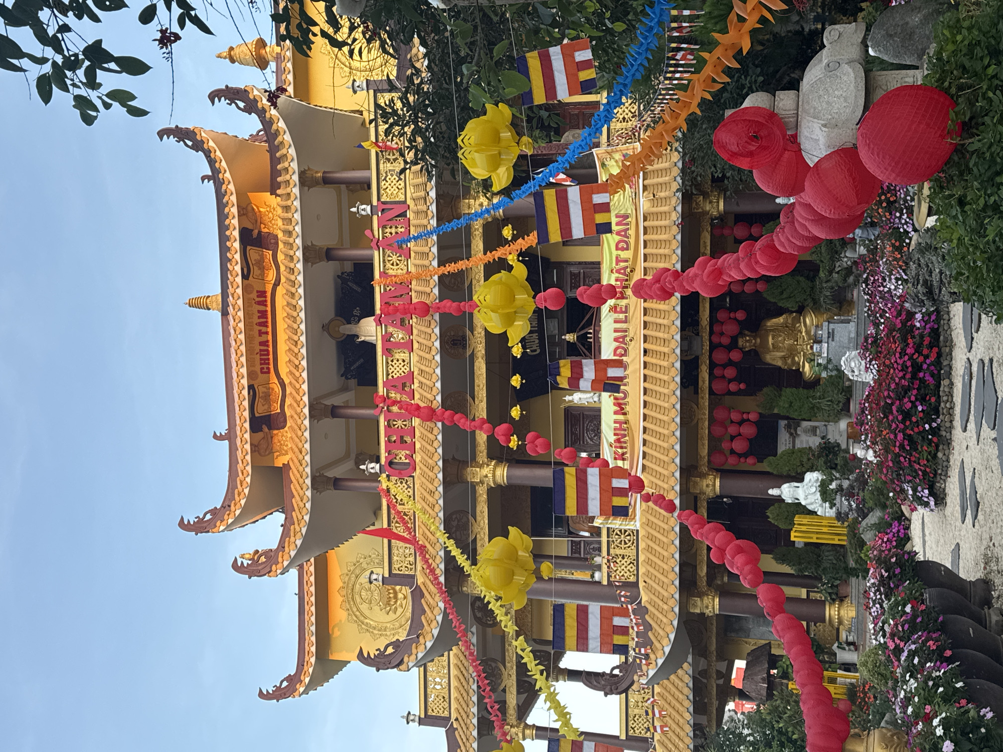 This image depicts the exterior of a Buddhist temple, likely in Da Lat, Vietnam, adorned with festive decorations. The temple features multi-tiered, ornate yellow roofs with intricate detailing and upturned eaves. Various prayer flags in red, yellow, blue, and white are hung along the building's facade, alongside colorful paper chains and large, yellow lotus-shaped lanterns. Red paper lanterns are also visible, both strung in garlands and hanging individually. A golden Buddha statue is prominently displayed within the temple complex, and a white Buddha statue is visible in the foreground garden area. The garden itself is lush with abundant flowers in shades of pink, purple, and white, planted in large dark pots and beds, with stone pathways winding through the greenery. The overall scene suggests a celebration or festival is being held at the temple. The sky above is clear and blue.