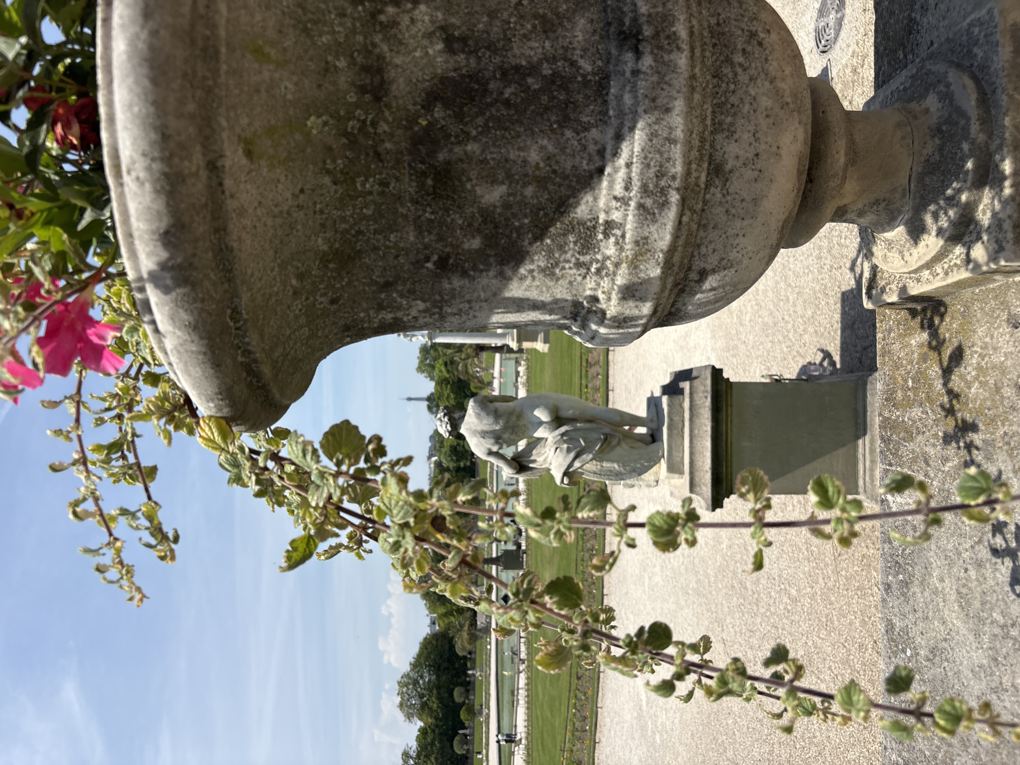 This is a medium shot, eye-level, outdoor photograph of a stone planter in the foreground. The planter is large, made of weathered gray stone, and has a curved, urn-like shape. It is filled with greenery and bright pink flowers, which are partially in focus. Some of the green foliage hangs down in front of the planter.
In the midground, to the left of the planter, is a stone statue of a nude figure from behind, draped with fabric. The statue stands on a pedestal. Beyond the statue, there is a park-like setting with a long body of water, manicured lawns, trees, and pathways. In the distance, a faint silhouette of the Eiffel Tower can be seen against a bright blue sky with some wispy clouds. The ground surface in the foreground and midground appears to be gravel or small stones. The overall scene is bright and sunny, suggesting a pleasant day in a public garden. This scene is likely the Luxembourg Gardens in Paris, France, due to the presence of the statue and the distant Eiffel Tower.