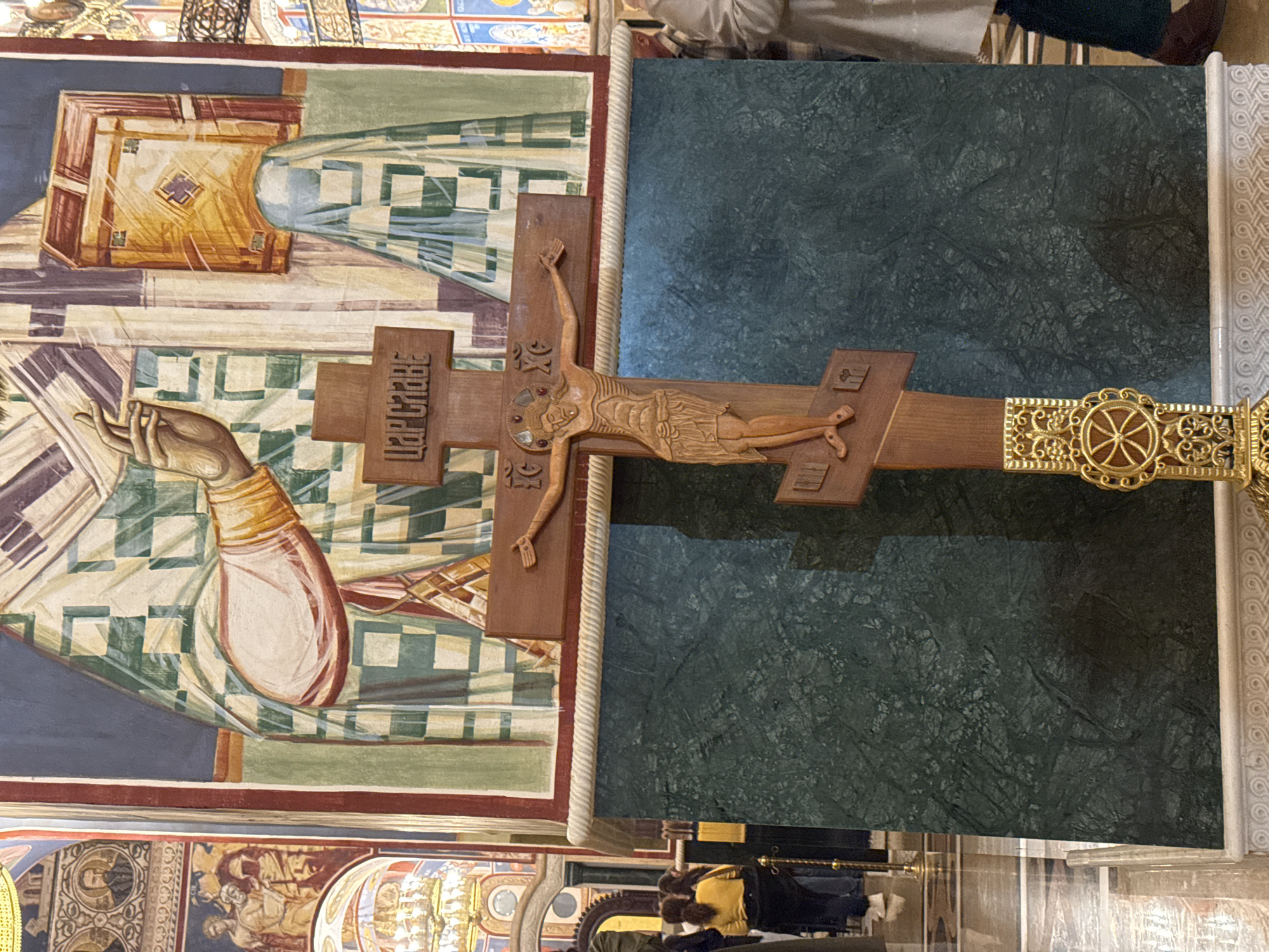 The image shows a crucifix positioned in front of a dark green marble-like panel. The crucifix appears to be made of wood, with a carved figure of Jesus Christ. Cyrillic text is inscribed on the cross. The cross has a gold-colored ornamental attachment on the right side. Behind the crucifix and panel is a painted icon depicting a figure holding a book or scroll. Below are other parts of an iconographic scene. The scene is in the city of Bar, Montenegro. People are faintly visible in the lower portion of the image.