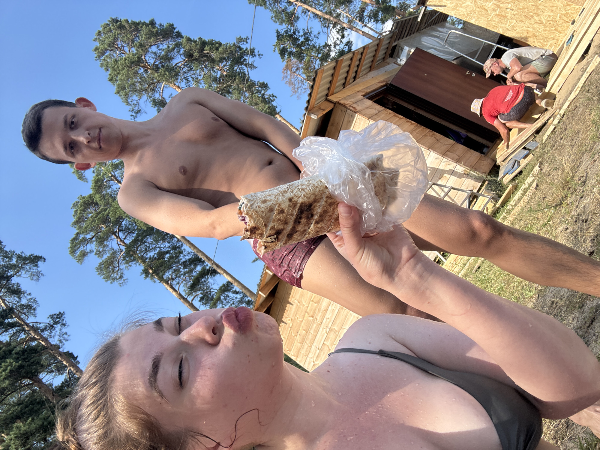 This media file captures a group of people in an outdoor setting, likely near a body of water given the wet skin and swimwear. The primary subjects are two young adults in the foreground, a woman and a man. The woman, in the immediate foreground, is looking towards the camera with her eyes closed and puckered lips, as if blowing a kiss. She is wearing a dark-colored bikini top and her skin appears to be glistening with water. She is holding a wrapped food item, possibly a shawarma or burrito, in plastic wrap, up towards the camera.
Behind her, a shirtless man is partially visible, leaning back. He is wearing burgundy swim trunks. In the background, two other men are engaged in some sort of construction or repair work on a wooden structure that resembles a small house or cabin. One man is wearing a red t-shirt and dark patterned shorts, and the other is in a grey t-shirt and light-colored shorts. They are both bent over, working on the structure.
The scene is set outdoors with a clear blue sky and tall pine trees visible. The ground is covered in dry grass and dirt. The lighting suggests it is daytime, likely late morning or afternoon, with bright sunlight casting shadows. The overall atmosphere appears relaxed and casual, with elements of leisure and a hint of ongoing activity in the background. The location context is Kurovskoye, Russia, which aligns with the natural environment depicted. No visible text is present in the image.