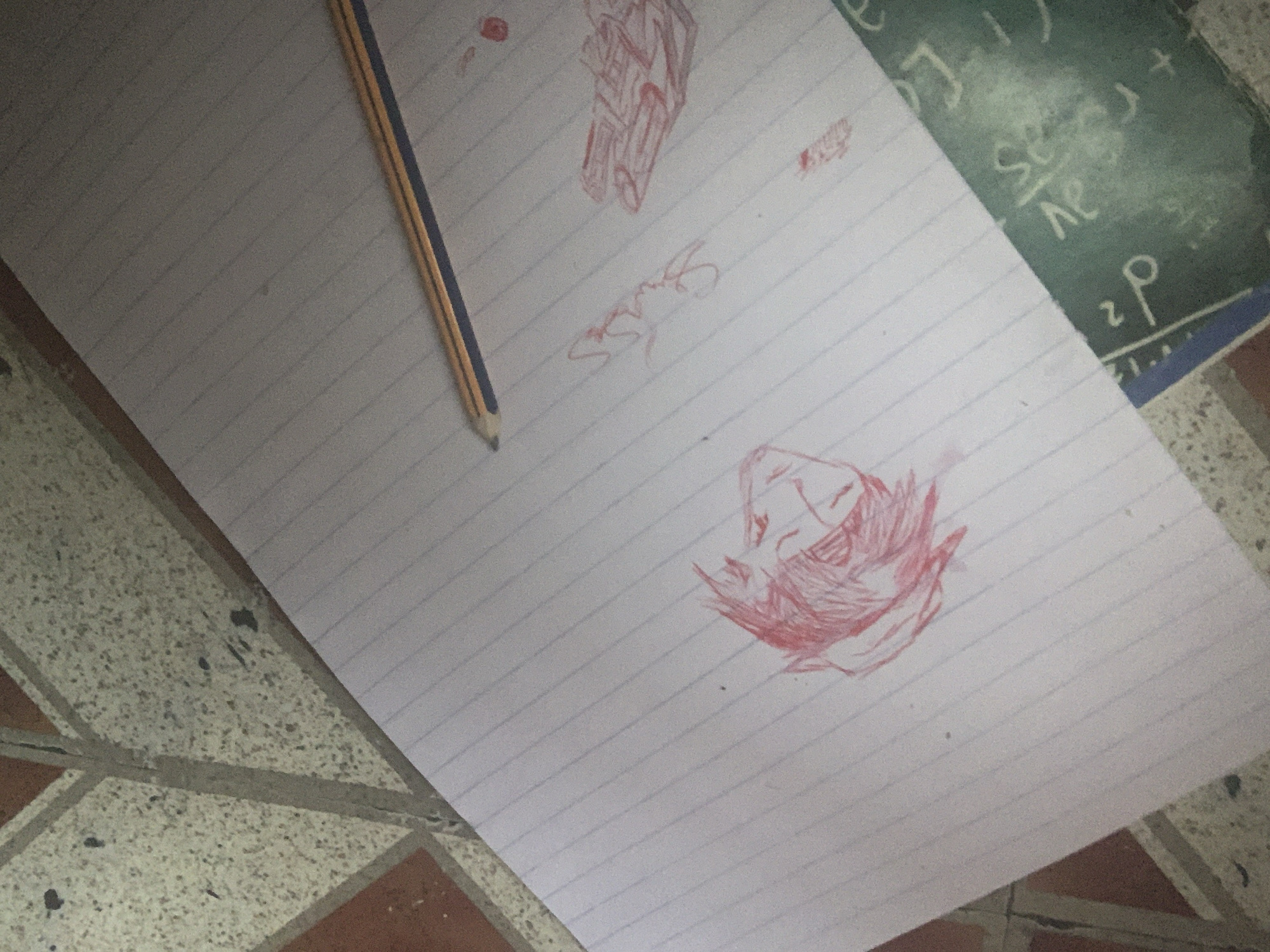 In Nkpor, Nigeria, a notebook with ruled lines is placed on a tiled floor. A pencil lies across the upper portion of the notebook. The notebook features several red crayon drawings. On the left side of the notebook, there is a sketch of a person's head with spiky hair. To the right of this sketch, there is some red scribbled text or a signature. Further to the right, there are red crayon drawings that appear to be of rectangular objects, possibly depicted as a brick pattern or a stack of items. A small red dot is also visible on the notebook. A green object with white writing, potentially a chalkboard or a slate with math equations, is positioned diagonally across the lower right corner of the notebook. The tiled floor consists of large, square tiles with a speckled pattern, separated by grout lines. Some dark spots are visible on the tiles.