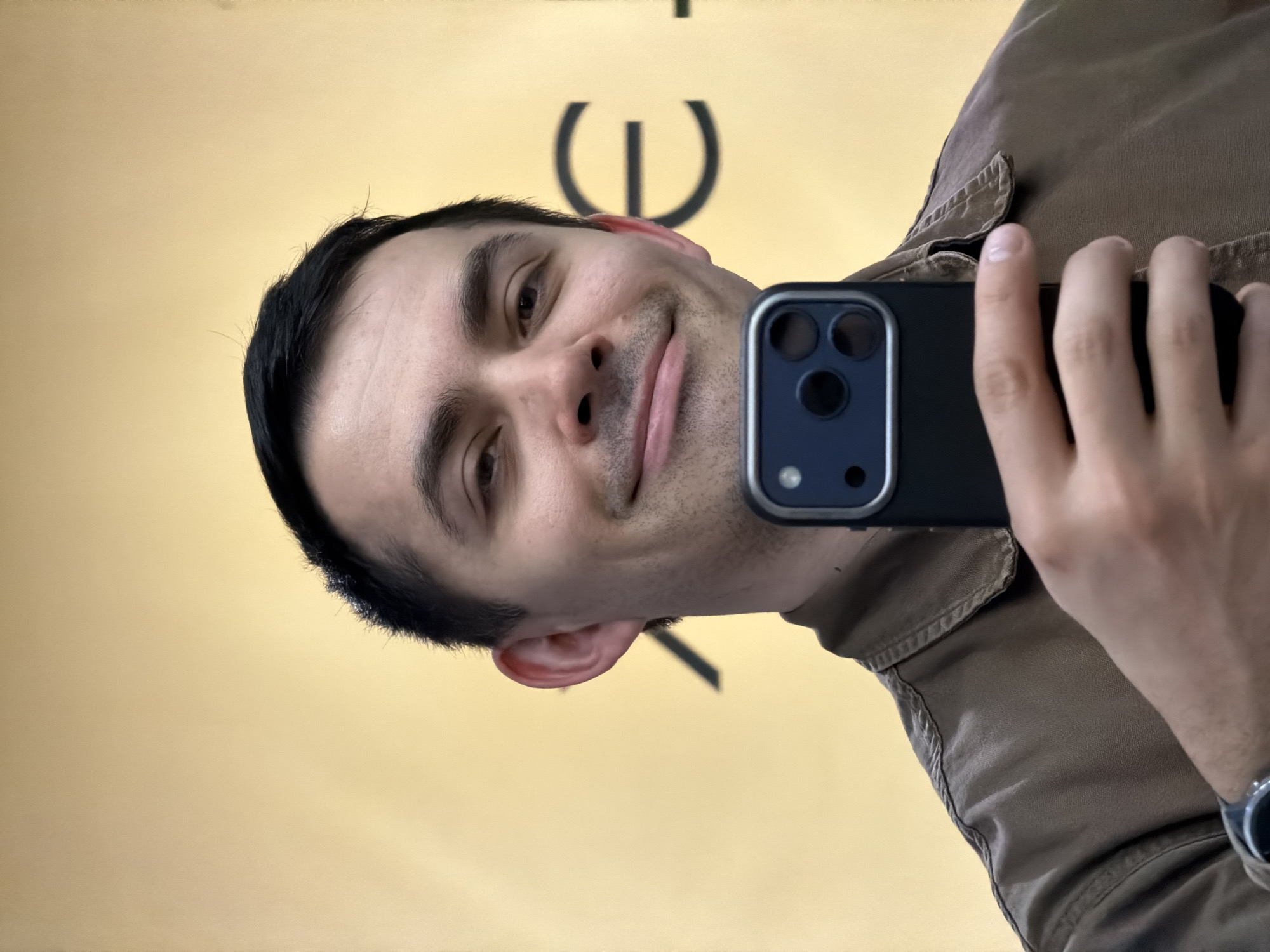 This image is a selfie-style reflection of a man in an indoor setting. The primary subject is a man with dark hair, a subtle smile, and short stubble, looking directly towards the camera. He is wearing a brown or olive-green collared shirt or jacket.
The man is holding a black smartphone with his right hand, positioning it in front of him. The phone clearly displays a triple-lens camera system on its back, characteristic of newer smartphone models.
In the background, behind the man's head and shoulder, a light yellow or beige wall is visible. On this wall, a large, dark, stylized letter "E" can be seen, appearing reversed due to the reflection. This letter is likely part of a word or brand name, possibly "ICE". The overall setting suggests an interior space, such as a store or a building with signage.
The image is captured at an angle, reinforcing the impression that it is a reflection, possibly from a mirror or window. The man's expression is calm and slightly amused. The provided location context for this media file is Izhevsk, Russia. No specific time of day or weather conditions are discernible from the image itself.