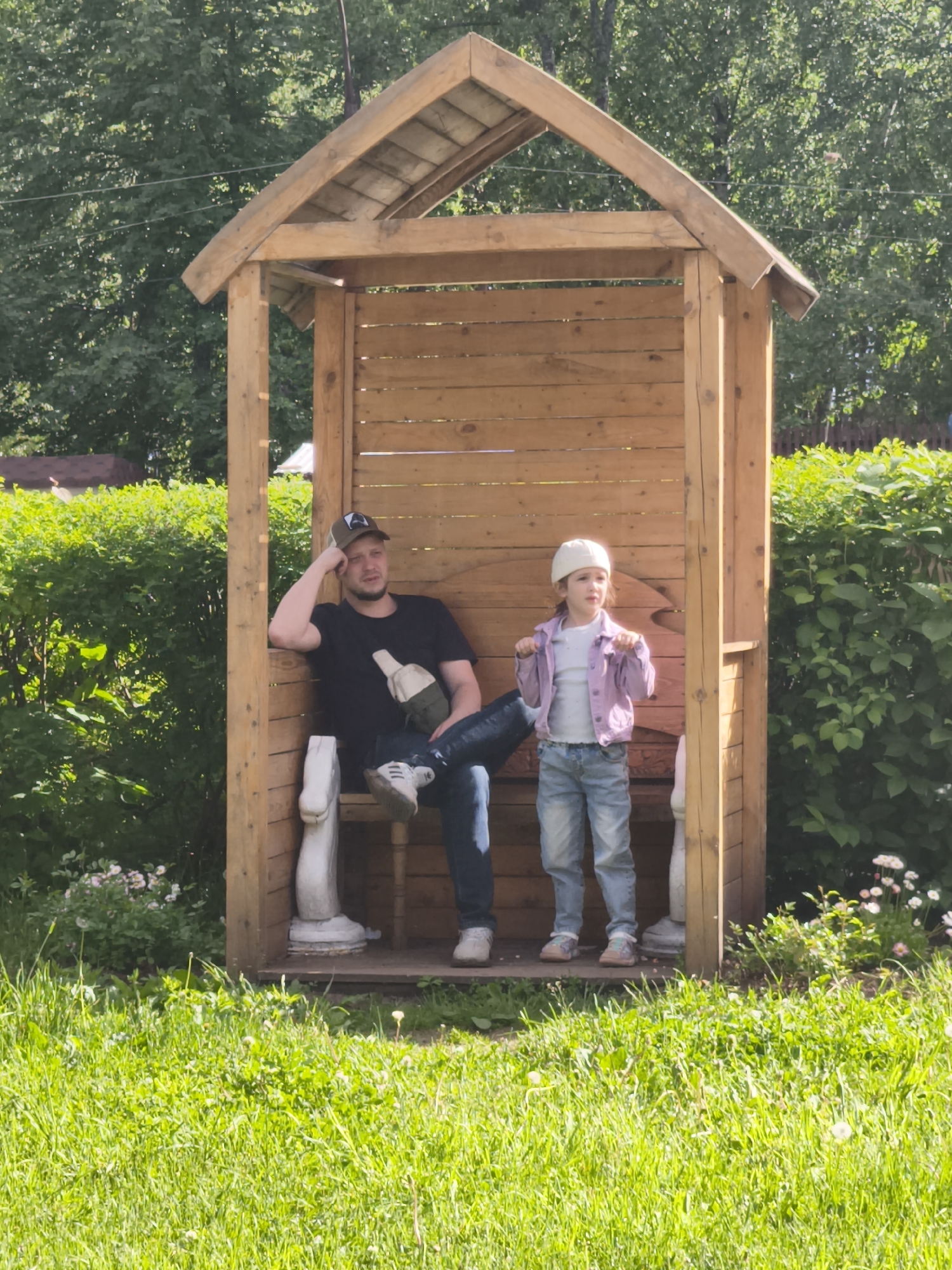 The image captures a scene in the city of Chaikovsky, Russia. A man wearing a baseball cap, black t-shirt, jeans, and white sneakers is seated inside a small, open wooden structure with a roof. He is resting his head on his hand and appears contemplative. A young girl, dressed in a white hat, light purple jacket, t-shirt, and jeans, stands beside him. Both are inside the wooden structure, which features decorative white statues on either side. The structure is set on a small wooden platform. The surrounding environment is a grassy area with green bushes and trees in the background. The lighting suggests it is daytime.