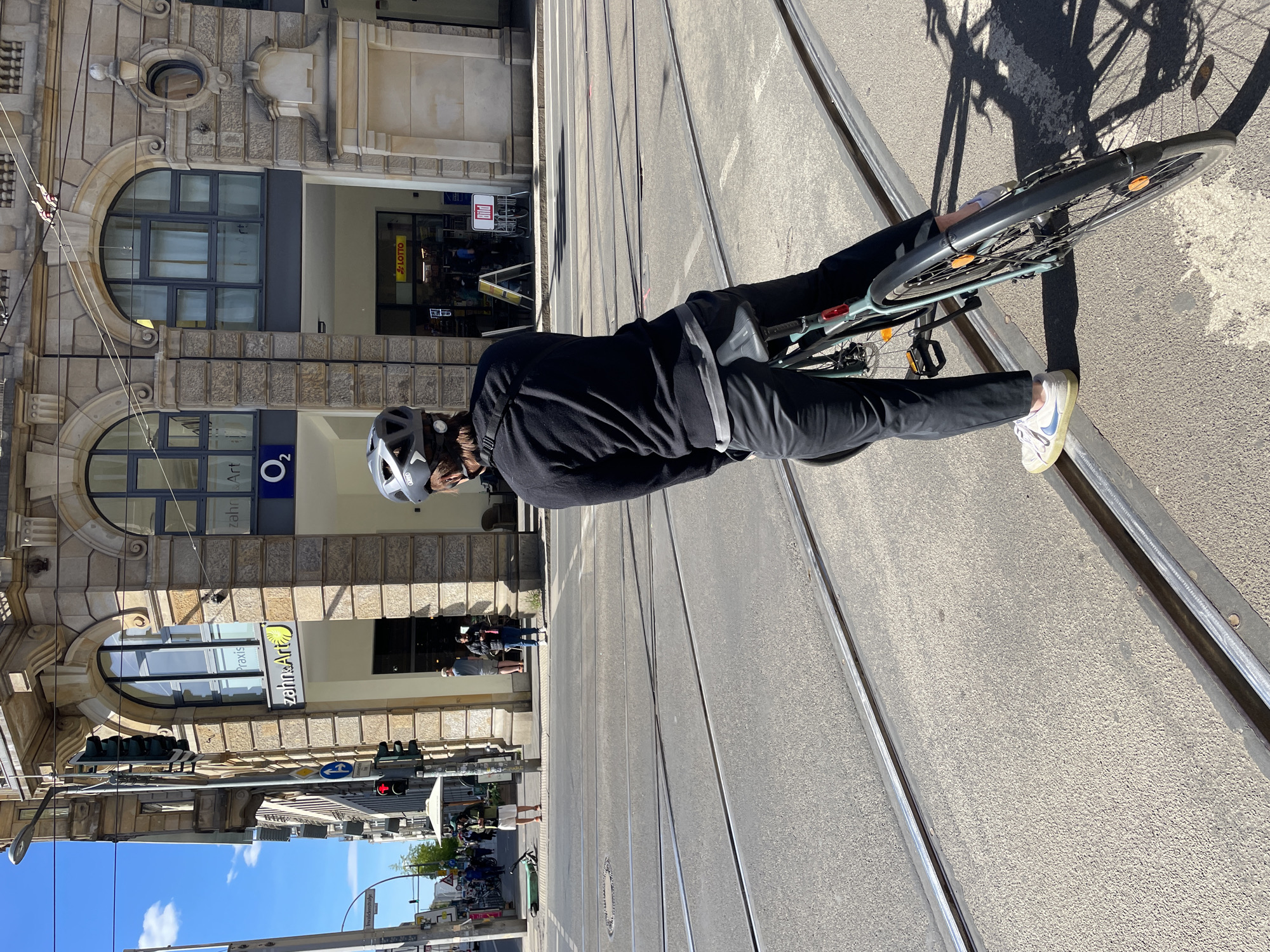 An urban street scene in Berlin, Germany, unfolds on a sunny day. The central subject is a person, seen from behind and slightly to the side, wearing a black long-sleeved top, black trousers, and light-colored sneakers with a Nike swoosh. They are also wearing a grey bicycle helmet and are standing with their light-colored bicycle on a street featuring multiple tram tracks. The bicycle's shadow is cast prominently on the asphalt to the right.
To the left, a grand stone building with ornate facades, arched windows, and decorative elements dominates the background. Several businesses are visible at street level. A prominent sign for "o2" (a mobile network provider) is seen above an entrance. Further down, another sign reads "Zahn & Art Praxis," indicating a dental practice. A "Lotto" sign is also visible, along with a "Bild" newspaper advertisement. Overhead, power lines for the tram run along the street.
In the mid-ground and distance, other pedestrians and street furniture like traffic lights (one showing red) and street lamps are visible. A street sign with "Invalidenstraße" partially readable confirms the Berlin location. The sky is clear blue with a few scattered clouds, and the bright sunlight casts distinct shadows, suggesting it is late morning or early afternoon. The overall impression is a typical day in a bustling European city.