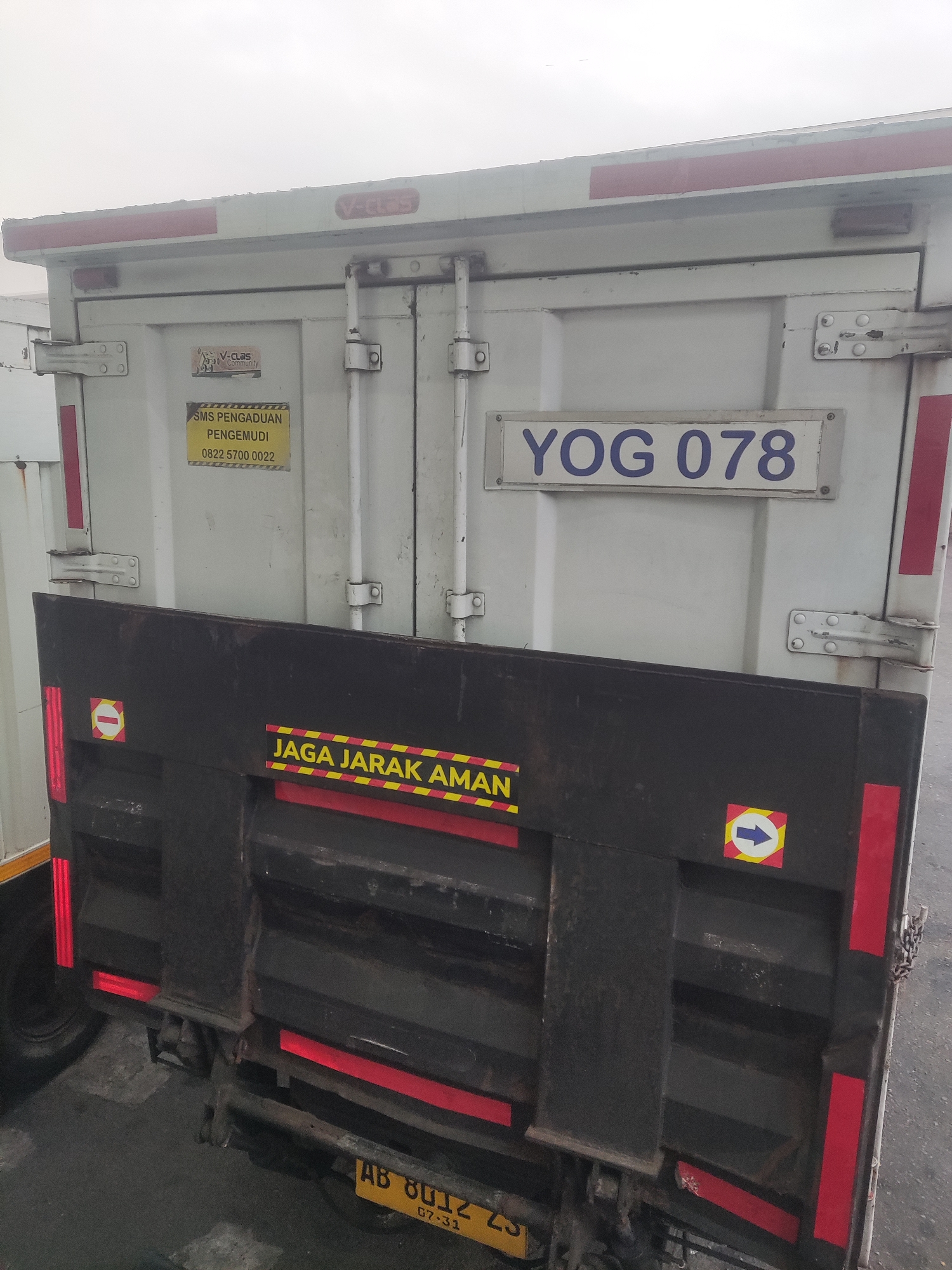 The image shows the rear of a white truck with the license plate YOG 078. A yellow sticker with the text "SMS PENGADUAN PENGEMUDI 0822 5700 0022" is visible on the truck's door. Below the door, a black flap with the text "JAGA JARAK AMAN" is present. The background is a slightly overcast sky. The truck is parked on a paved surface. The lower portion of the image shows a yellow license plate that reads "AB 8012 ZS 07-31". The overall scene is consistent with a vehicle parked in an outdoor location, possibly a loading dock or street in Trihanggo, Indonesia.