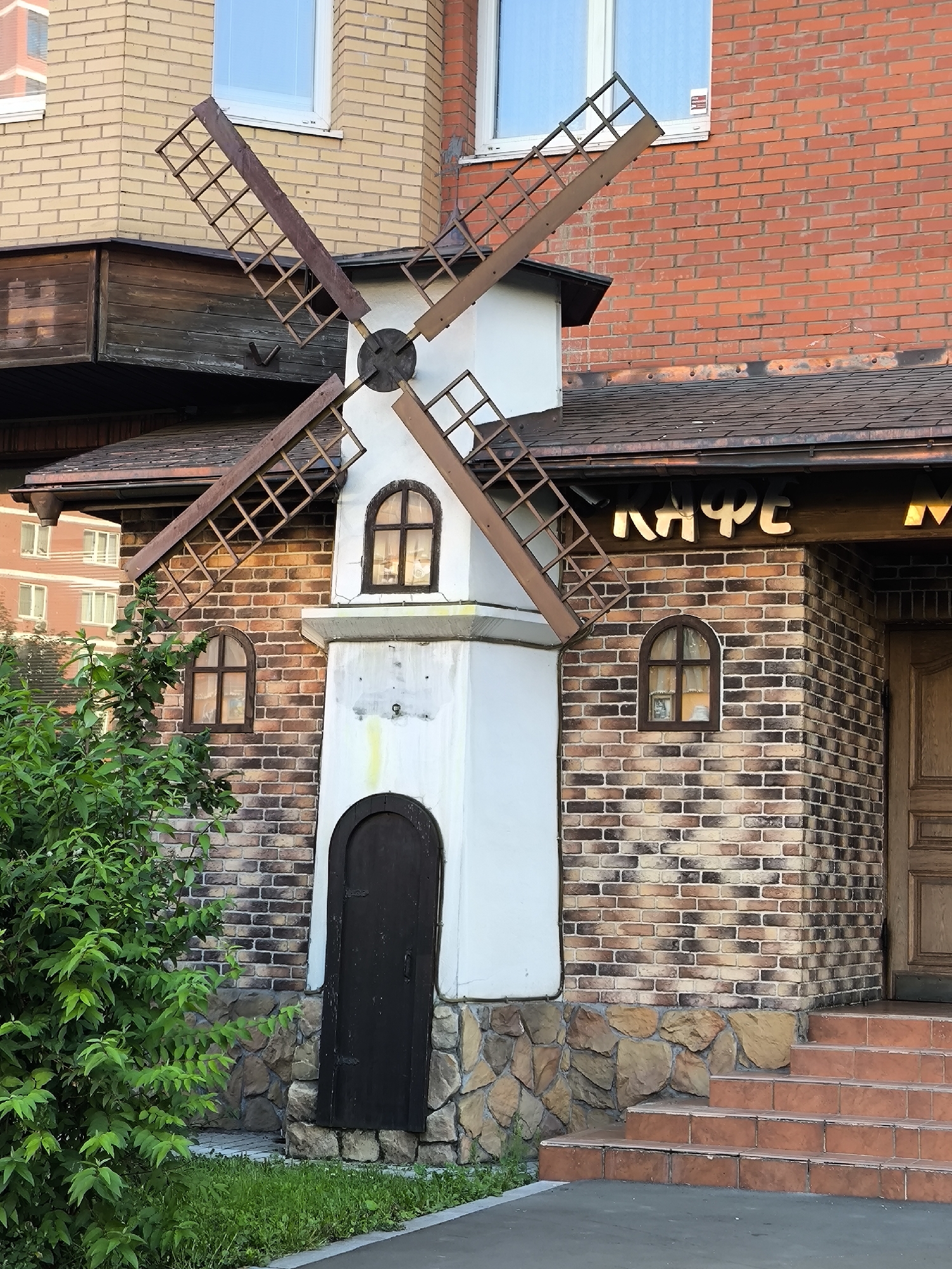 An outdoor, eye-level, medium shot captures a decorative windmill structure attached to a brick building, possibly a cafe, in an urban setting. The windmill, a quaint design with wooden sails, is positioned in front of the brick facade of a building. The building features a dark-tiled roof, arched windows, and a prominent sign with Cyrillic letters that read "КАФЕ" (Cafe). The lower portion of the building is adorned with stonework, leading up to a set of reddish-brown tiled steps. To the left, a lush green bush with dense foliage partially obscures the view of the building. The background reveals glimpses of other buildings, suggesting a city environment. The lighting indicates it is daytime, with the sun casting subtle shadows. No people are visible in the frame.