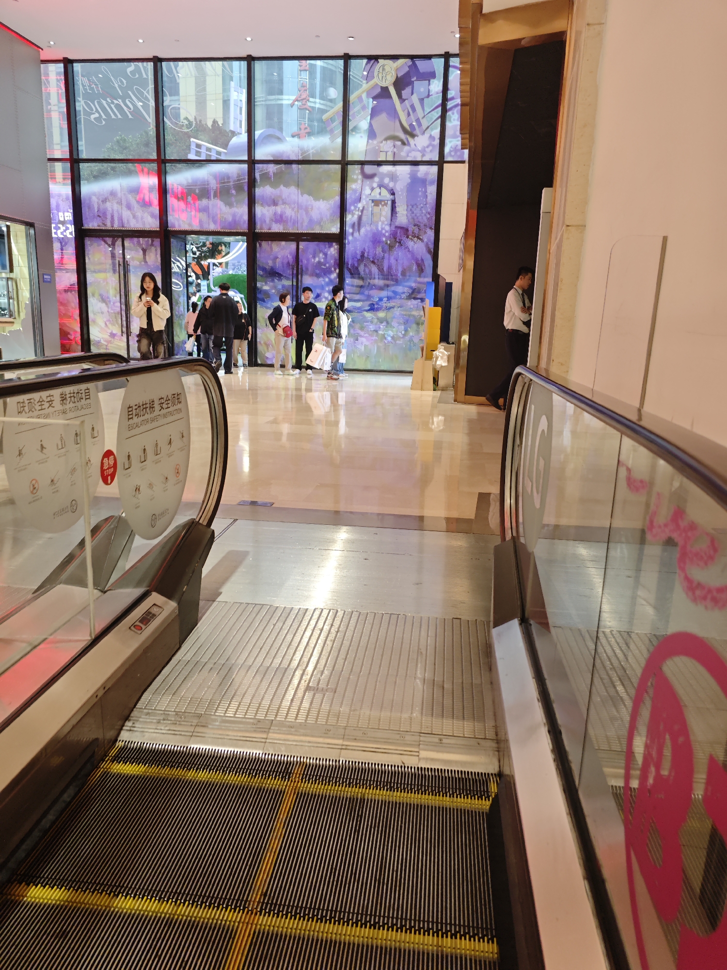 A ground-level, indoor shot depicts the interior of a building with a wide-open entrance area, most likely a shopping mall or commercial center, in Yuzhong District, China. The foreground features a non-operational escalator, with its treads showing black lines and yellow highlights. The glass panels on either side of the escalator have safety instructions and signs.
Beyond the escalator, a well-lit, glossy-tiled floor extends towards a large wall of windows and doors. The windows showcase an artistic display featuring what seems to be a landscape with lavender fields and stylized windmills. Several people are standing or walking near the entrance, including individuals dressed in casual attire. A man in a white shirt is standing near the side wall of the building. The environment is clean and modern, suggesting a contemporary retail or commercial space.