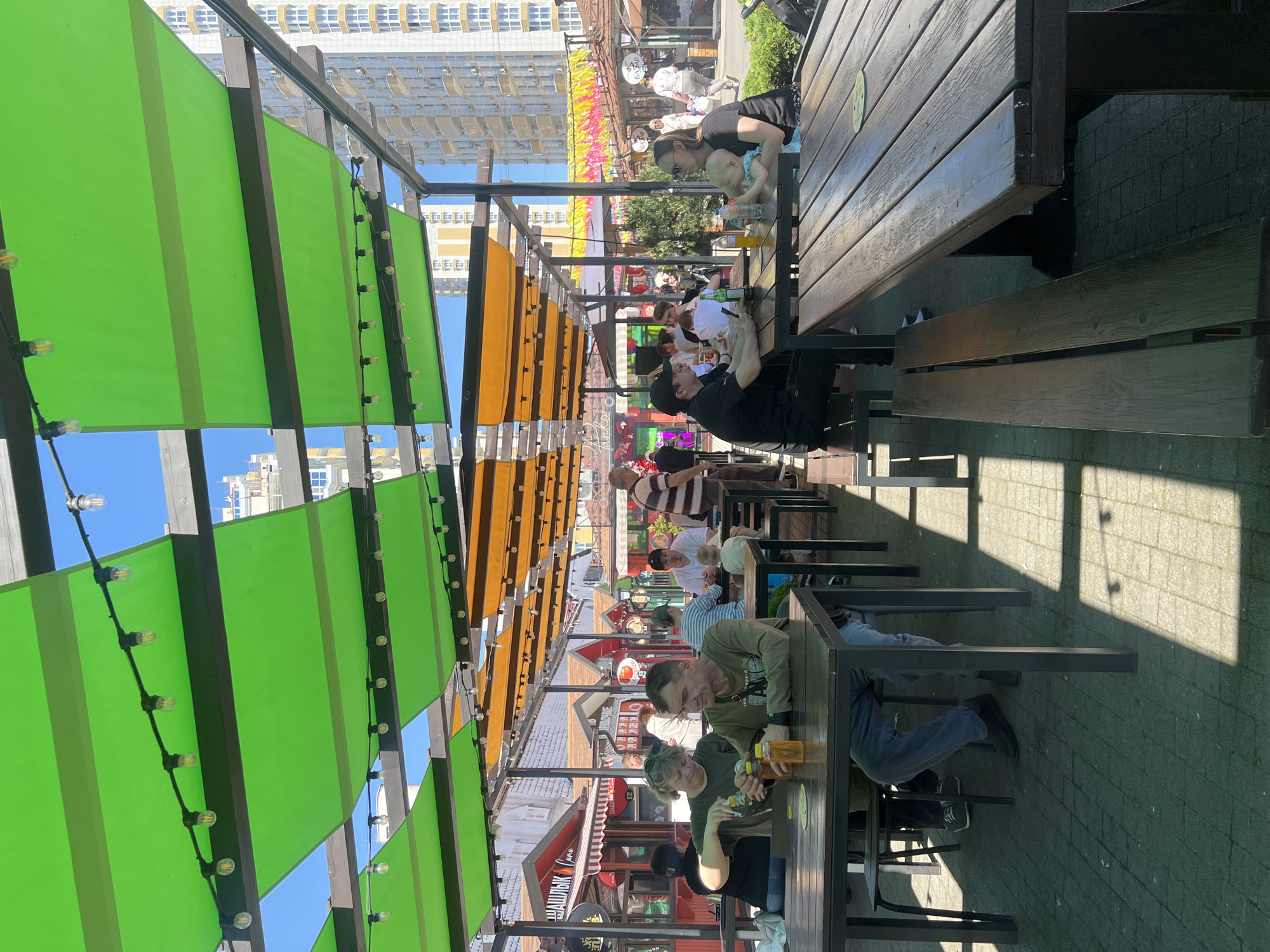 The image depicts an outdoor seating area of a restaurant or café. There are several wooden tables with benches arranged under a canopy structure. The canopy features alternating green and orange fabric panels, strung with small decorative light bulbs. People are seated at the tables, some appear to be eating or drinking. A tall building is visible in the background. This location is likely Krasnodar, Russia.