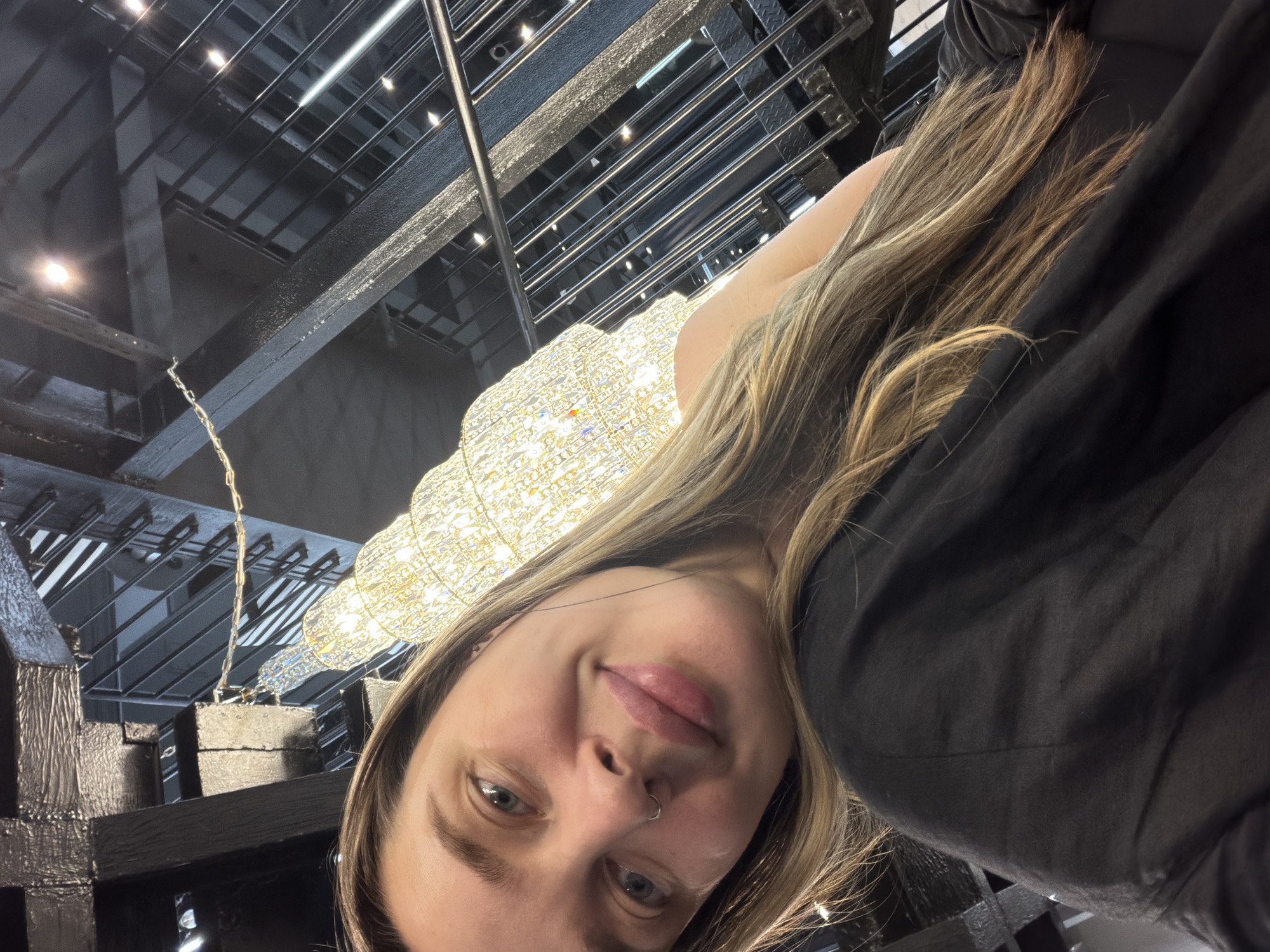 A young woman is posing for a selfie in what appears to be a modern architectural space. She has light brown hair, fair skin, and a nose piercing. She is wearing a dark, flowing top or dress. Behind her is a large, intricate chandelier or light fixture composed of many small, sparkling elements. The ceiling and the surrounding structure feature a dark, grid-like pattern with exposed beams and metalwork, suggesting an industrial or contemporary design. The specific location in Moscow, Russia, is not discernible from the given image.