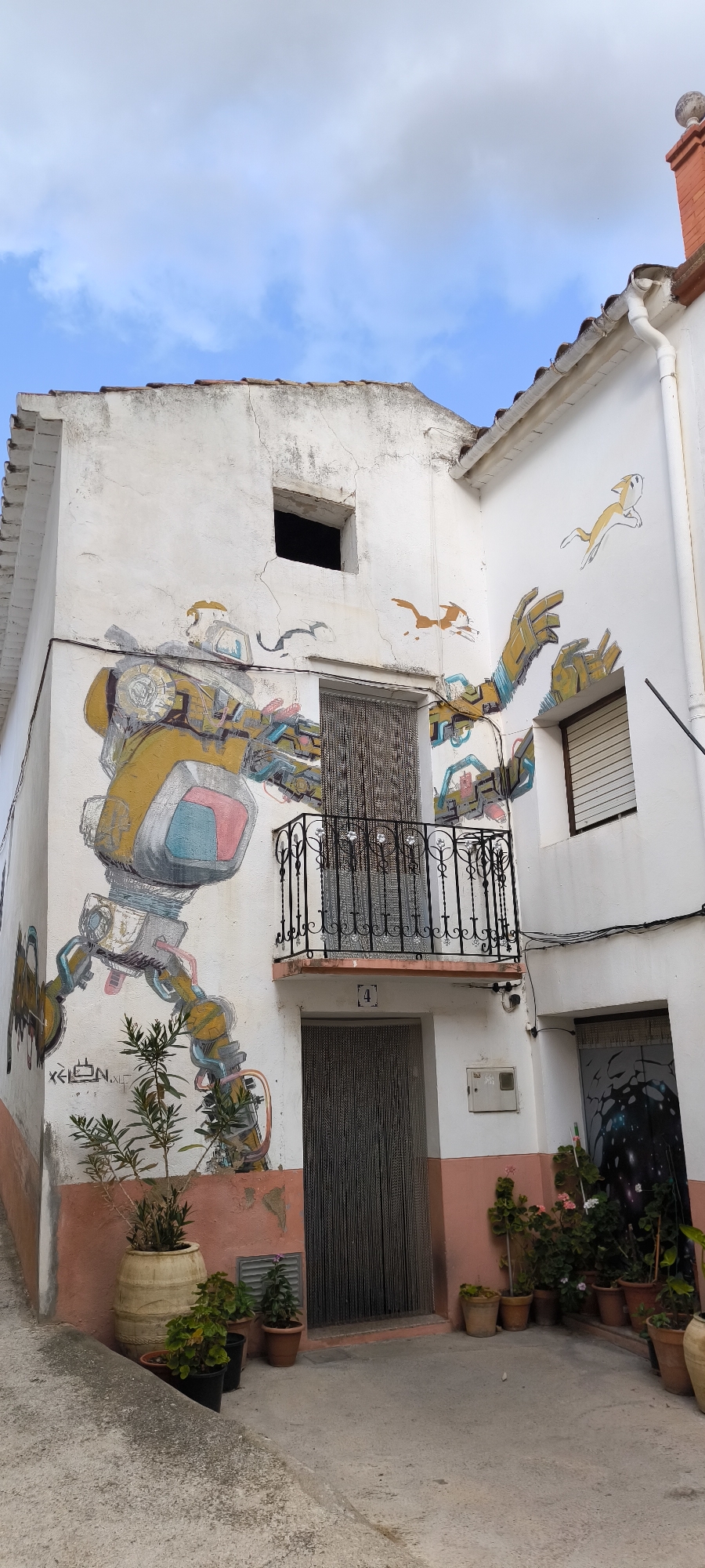 The image depicts a street scene in Fanzara, Spain, featuring a building adorned with vibrant murals. On the left side of the building, a large, colorful mural of a robot is prominently displayed, with intricate mechanical details. To the right of the robot, there are smaller murals of stylized animals, appearing to be leaping or running. The building itself is white with pink trim along the base. A balcony with black wrought-iron railings is situated above a doorway, which is covered by a beaded curtain. Another doorway, also covered by a beaded curtain, is visible to the right. Potted plants are arranged at the base of the building, adding a touch of greenery to the scene. The sky above is partly cloudy with a blue hue. The street in front of the building is made of concrete. The overall impression is one of an urban environment enhanced by public art.