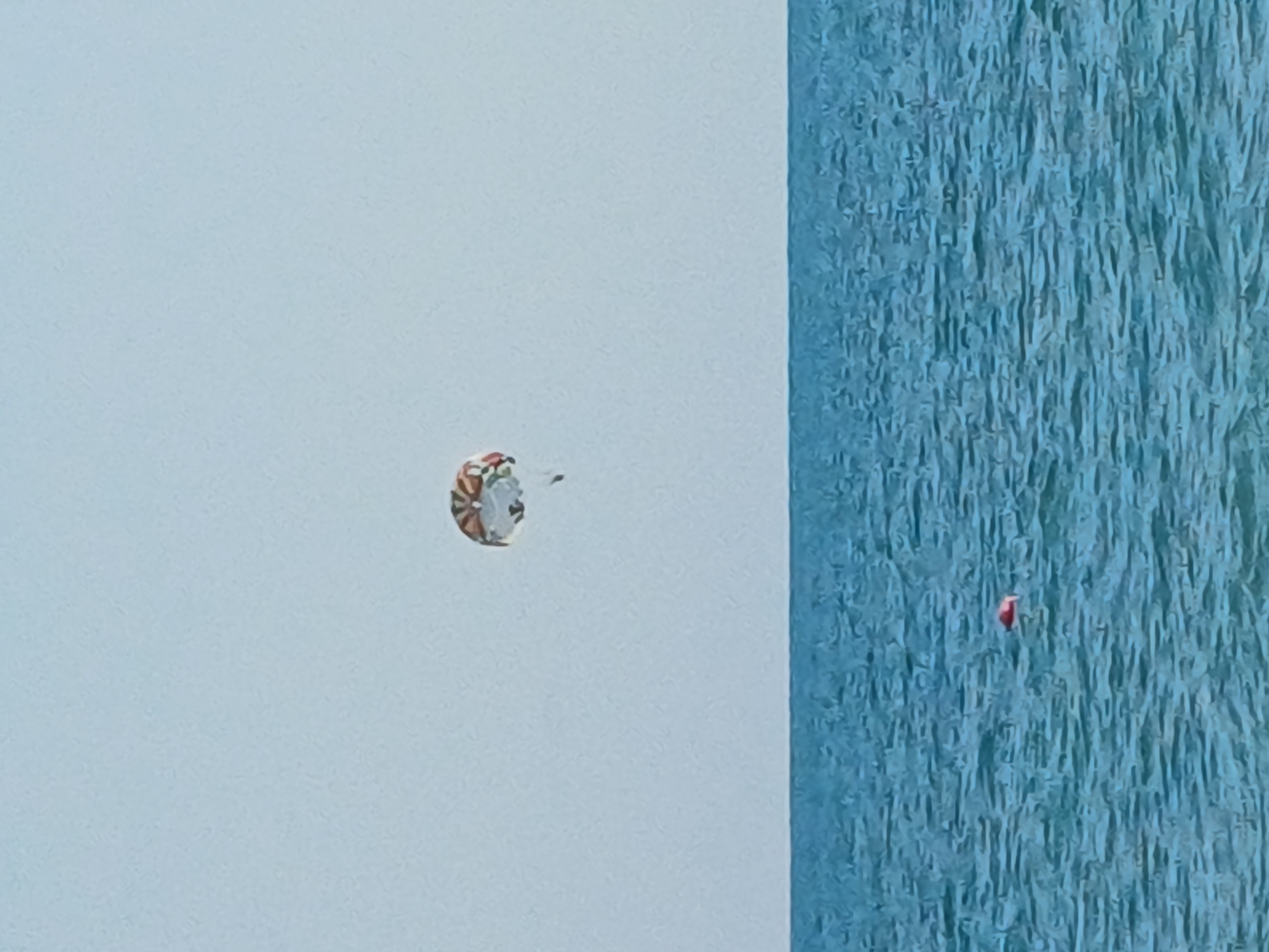 The image shows a paraglider in mid-air over a blue, textured surface that appears to be water. The paraglider is small in the frame and is multicolored with a patterned canopy, likely designed for visibility. It is positioned against a pale blue sky, which occupies the left half of the image. The right half of the image is dominated by the textured blue surface, which exhibits vertical striations, suggesting waves or ripples on the water.
A small, red object is visible in the water, to the right of the paraglider. Its nature is unclear from the image, but it could be another paraglider's chute, a marker buoy, or debris.
The scene appears to be set during daylight, given the bright sky. There are no strong indications of specific weather conditions, other than the clear sky. The textured water suggests a body of water, possibly the sea or a large lake. The mention of Sochi, Russia, places this scene in a coastal resort city known for its beaches and outdoor recreational activities, making paragliding a plausible activity. The image captures a moment of flight and recreational activity, with the vastness of the sky and water emphasizing the freedom and scale of the paraglider's journey. No text is visible in the image.