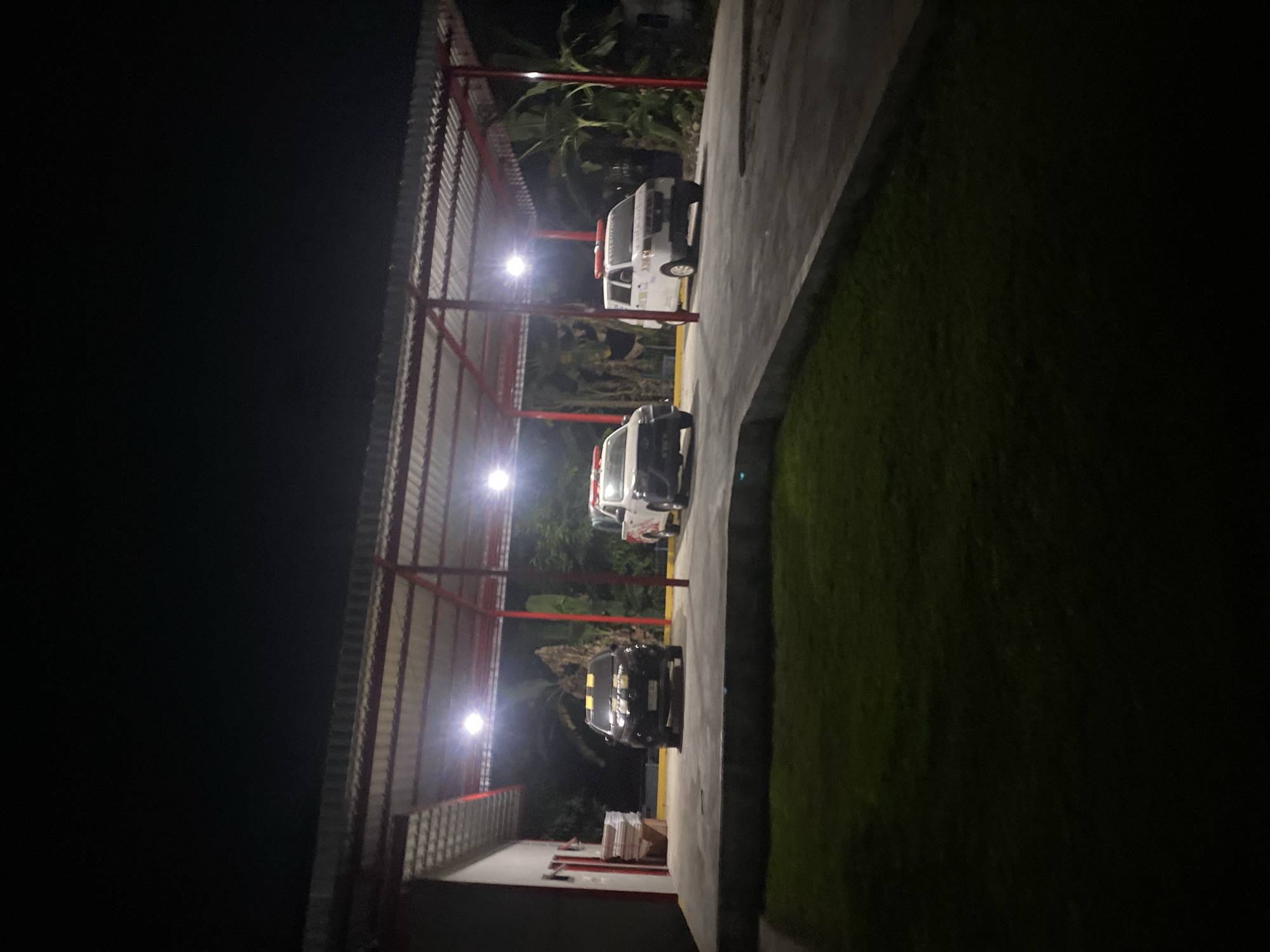 This is a vertically oriented photograph taken at night. The scene is an outdoor covered parking area with a corrugated metal roof supported by red metal posts. Several vehicles are parked underneath the shelter.
In the foreground, a dark-colored sedan is parked. Its headlights are on, casting a bright reflection on the concrete surface. Behind the sedan, another vehicle, possibly a white van or utility vehicle with red accents and what appear to be emergency lights on top, is visible. Further back, another white vehicle is parked. The vehicles are parked in a row, under the shelter.
To the right of the parking area, there is a grassy area, which is dimly lit by the overhead lights of the shelter. Beyond the shelter, to the left and rear, there are trees and vegetation, silhouetted against the dark night sky. The overall impression is one of a functional, possibly service-related parking area, operating at night. There are no people visible in the image. The lighting suggests artificial illumination from overhead fixtures within the covered area.
Some text is visible on the white vehicle in the middle ground; it appears to be lettering, but is not clearly legible due to the distance and lighting. The time of day is clearly night. There are no visible weather cues. The presence of vehicles, including what may be emergency or service vehicles, and the sheltered parking suggests a location like a depot, station, or a business premises. The mention of Banjarbaru, Indonesia provides geographical context.