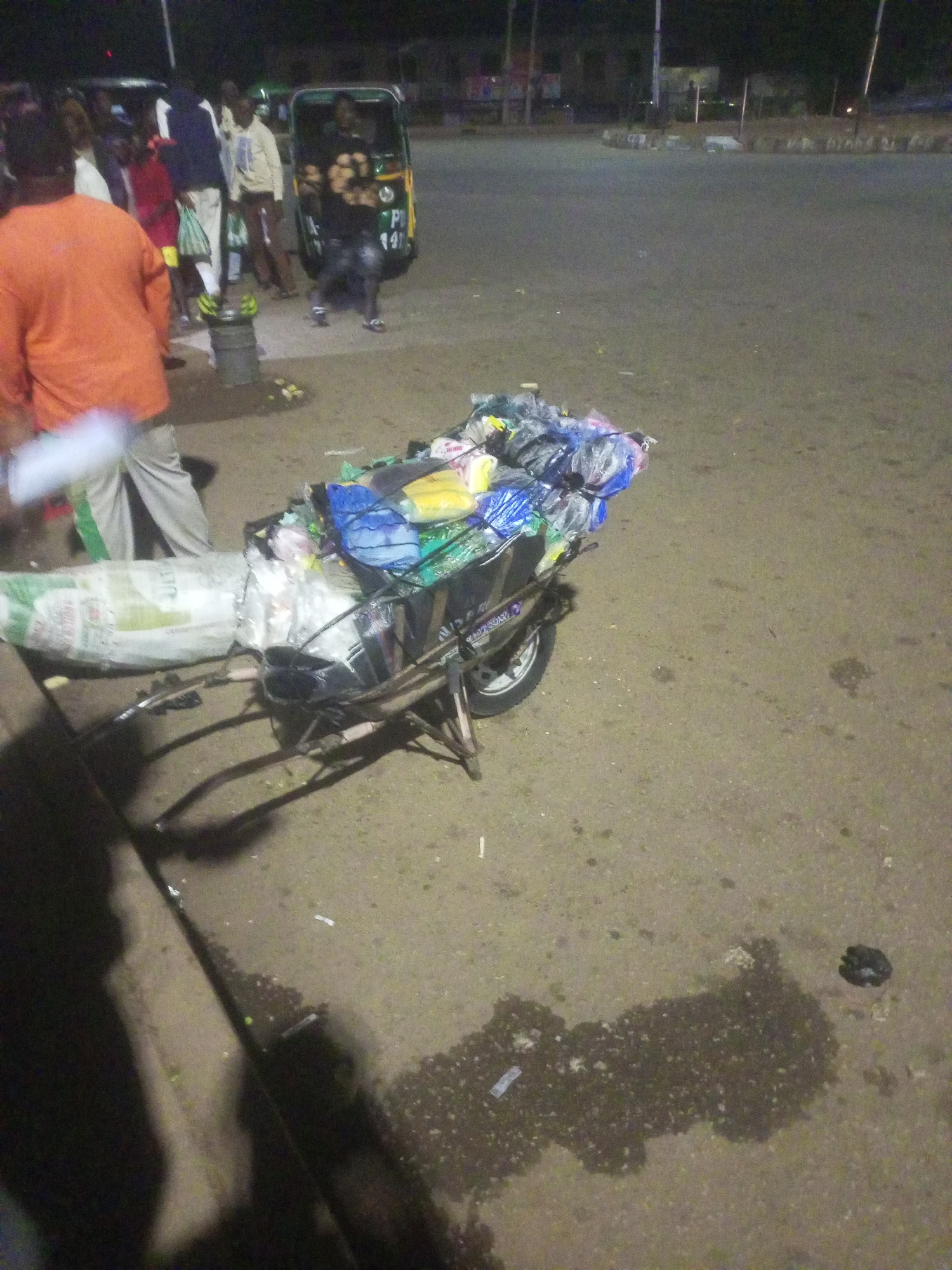 The image captures a nighttime street scene in Bukuru, Nigeria, characterized by dim lighting and artificial light sources in the background.
In the foreground, a two-wheeled handcart is heavily loaded with various items, predominantly merchandise contained within colorful plastic bags (blue, yellow, black, white, and green). A large white sack, possibly containing bulk goods with the faint text "ULTI" visible, is secured to the front of the cart. The cart's single wheel is visible on the right.
Several people are present in the mid-ground and background. On the far left, a person wearing an orange shirt and light-colored pants stands with their back to the camera. Other individuals are gathered in the background, some near a green and black auto-rickshaw (tuk-tuk) which displays partial text "P" and "147" on its side. A gray bucket, topped with yellow or green items resembling bananas, is placed on the ground among the people.
The ground is paved, appearing somewhat dusty and marked with several wet patches. A raised curb or sidewalk edge is visible along the left side. The scene suggests an active late-night market or street vending environment. No specific emotions are discernible among the individuals.