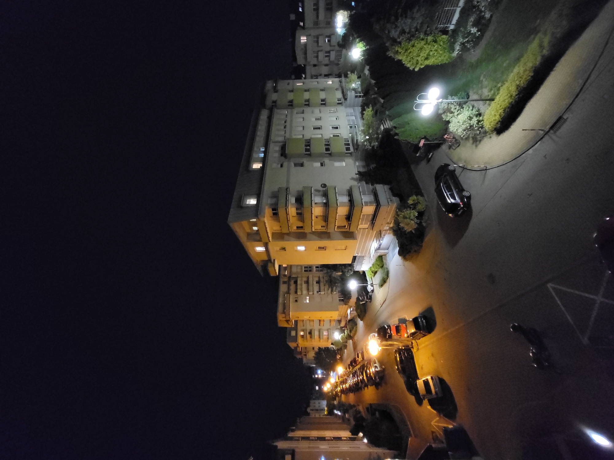 This image shows a nighttime view of a city street. The street is lined with parked cars and illuminated by streetlights. A tall building stands on one side of the street, with a dark sky visible to the left. A parking lot is on the right.