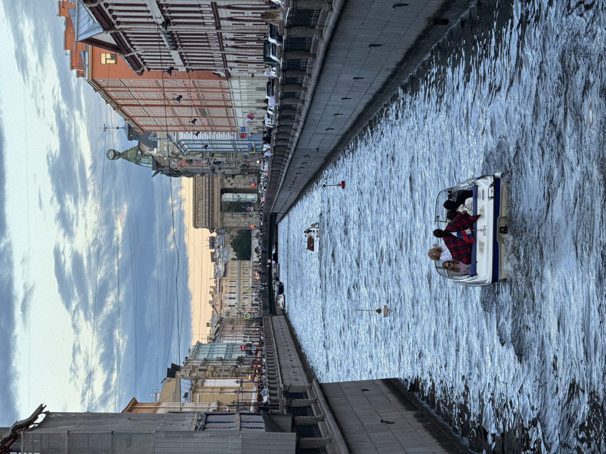 The image shows a view down a canal in Saint Petersburg, Russia, with ornate buildings lining both sides. A small white motorboat with four people on board is moving away from the viewer towards a bridge in the distance. The canal is bordered by stone embankments and a walkway with railings. Several other boats are visible further down the canal, and people can be seen walking along the embankment. The sky is cloudy with hints of sunset. In the distance, the Kazan Cathedral is visible with its semicircular colonnade and dome.