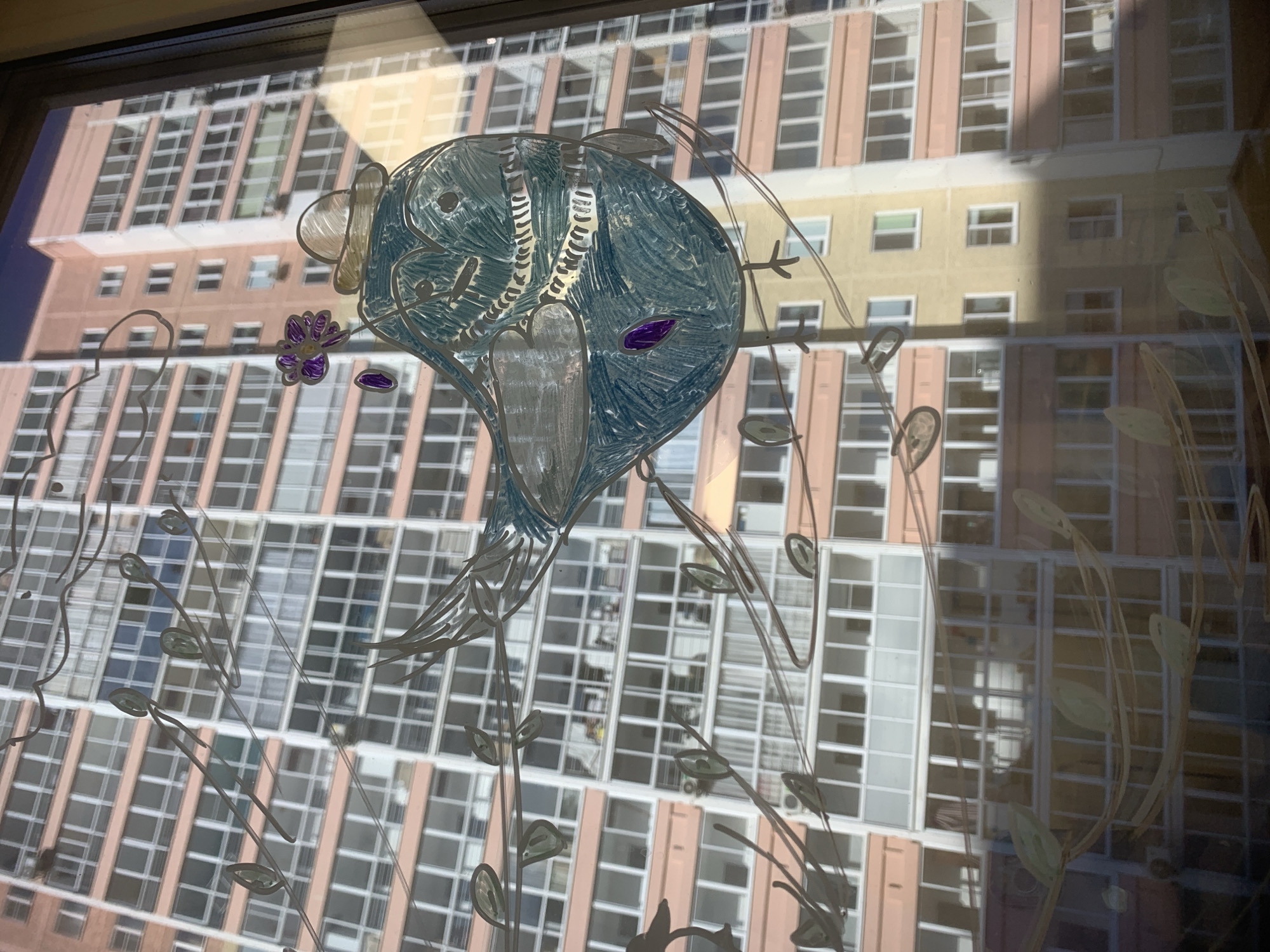 The image shows a window with drawings on it, depicting a blue bird-like figure with a flower and several stems with leaves. The background is a tall building with many windows. The location is not identifiable as Izhevsk, Russia based on the image alone.
