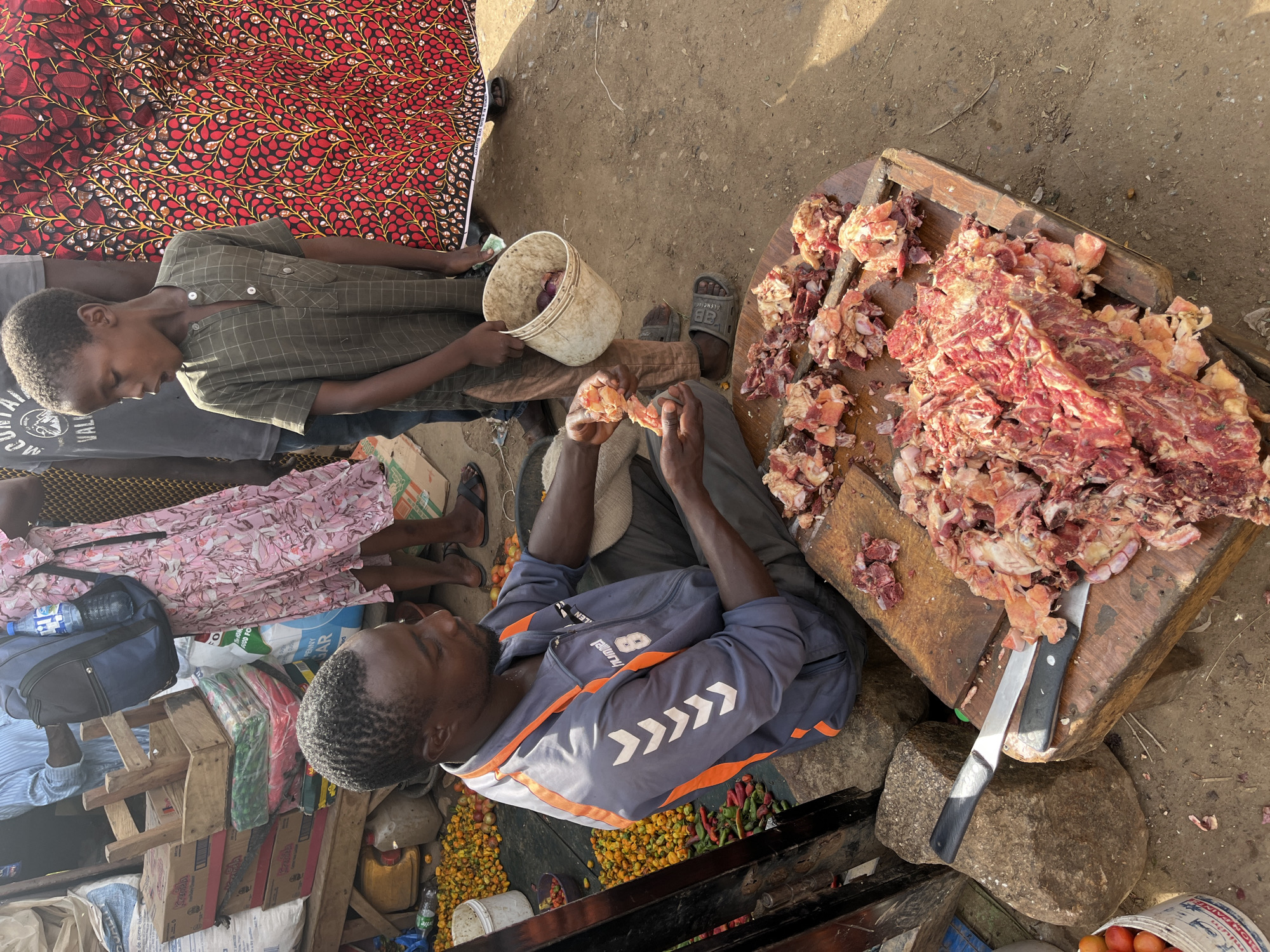 This media file depicts an outdoor market scene in Rafin Par, Nigeria. The primary subjects are two individuals, likely a young boy and a man, engaged in preparing meat. The boy, wearing a green checkered shirt, holds a white bucket containing red onions and appears to be assisting. The man, in a blue and orange jacket with "hummel" and "82" text visible, is seated and working with a large pile of raw meat on a wooden chopping board. A knife is present on the board.
The setting is a dusty ground, typical of an outdoor market. To the left of the frame, a woman in a pink floral dress is partially visible. Various objects are scattered around, including bags, boxes, a water bottle, and a yellow jerrycan, suggesting a bustling marketplace. Piles of colorful peppers and tomatoes are also visible, indicating the sale of produce.
The image appears to be taken during daylight hours, with sunlight casting shadows on the ground. The overall atmosphere seems focused on the task at hand, with no strong emotional expressions readily apparent. The visible text includes brand names like "hummel" and "ALENCIAG," as well as what appears to be partial words or phrases from other products or signage. The presence of fresh meat and produce points to a food market setting.