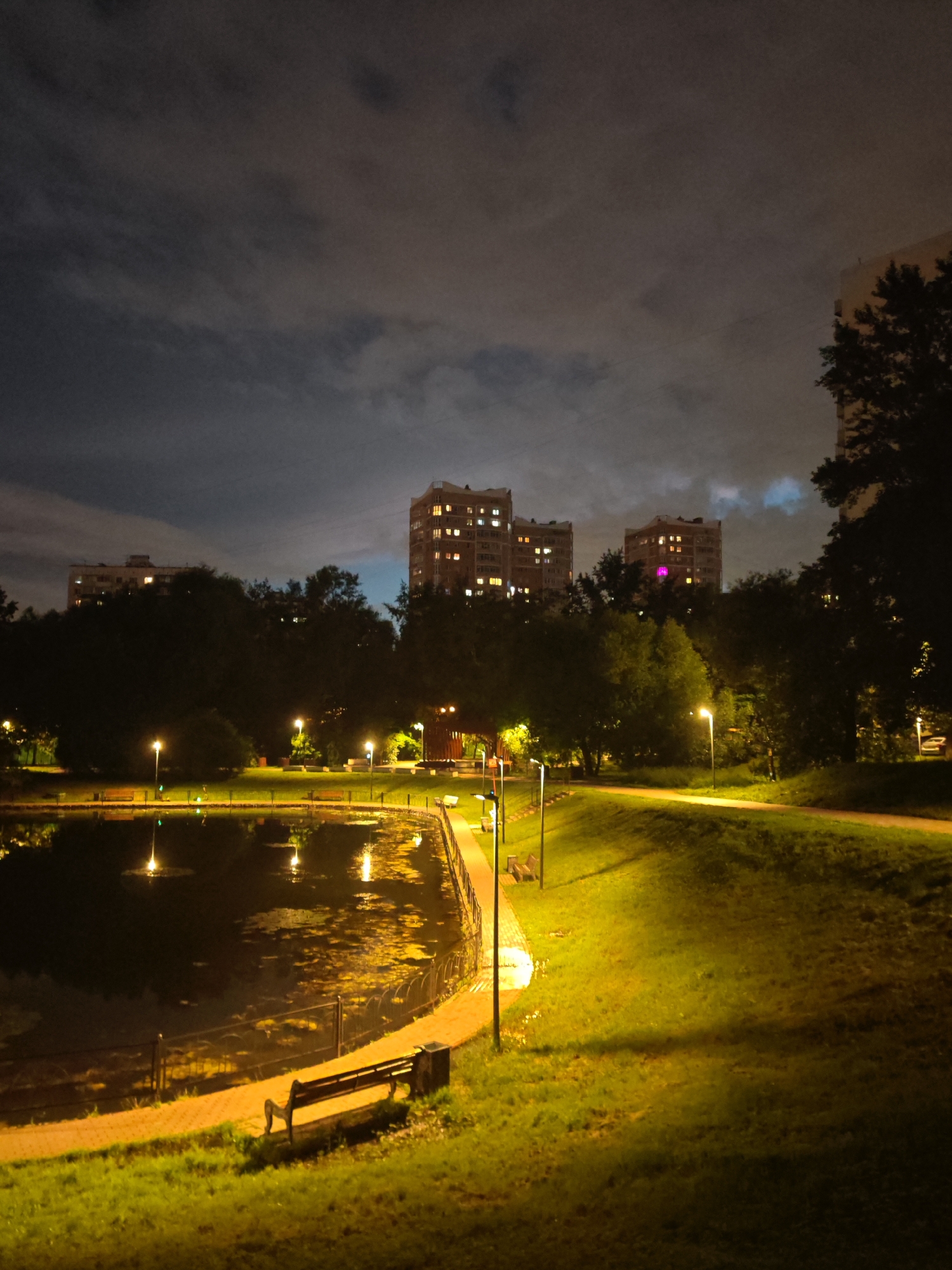 The image depicts a tranquil park scene at night in the city of Unknown, Unknown. A calm body of water, likely a pond or lake, reflects the ambient light from the surrounding streetlights and illuminated buildings. The water's surface is dotted with patches of vegetation. A paved path encircles the water, bordered by a metal railing. Several park benches are visible along the path, one in the foreground with a waste bin next to it. The surrounding area is covered in green grass and scattered trees. In the background, several multi-story buildings are lit up, their windows glowing with interior light. The sky above is cloudy and dark, with some areas of lighter cloud cover illuminated from below by artificial lights. The overall atmosphere is serene and peaceful, typical of a well-maintained urban park after dark. There are no people visible in the image.