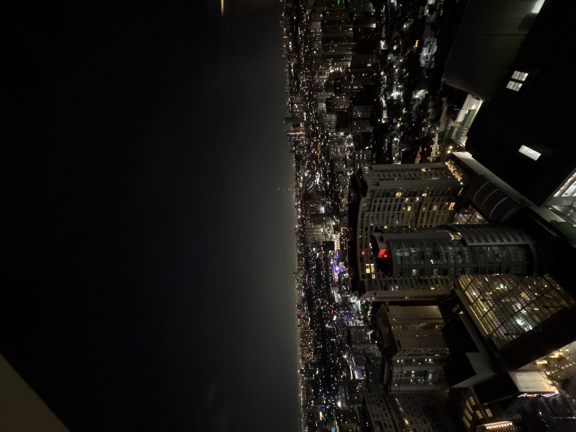 The image presents a night view over Jesús del Monte, Mexico. On the right side, a dense cityscape is visible, punctuated by illuminated buildings. Building styles appear modern, including curved glass facades and high-rise structures. Many lights fill the scene. To the left is a dark expanse, likely a body of water or open space, with a faint horizon line. The image is rotated 90 degrees clockwise.
